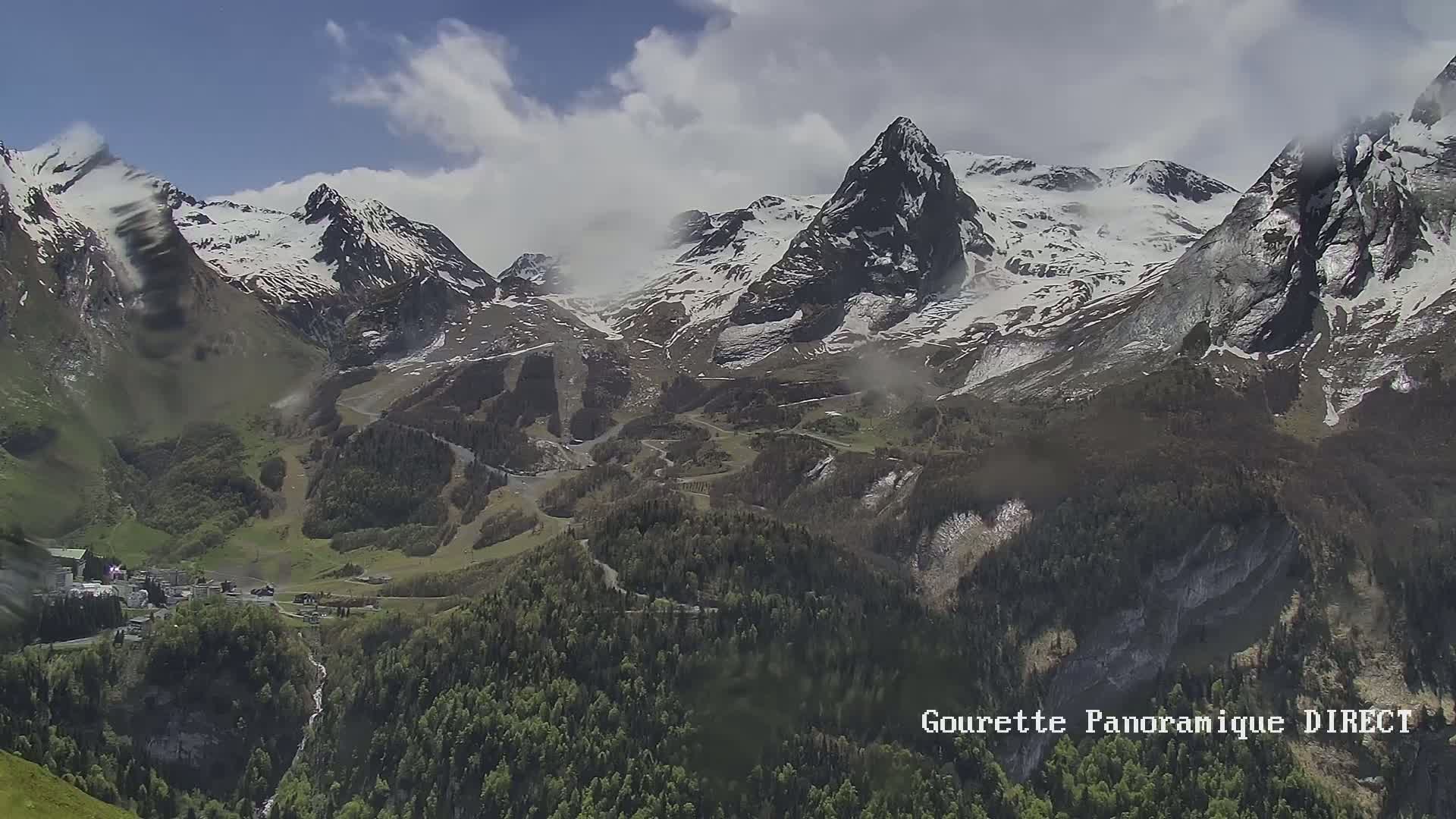A partly cloudy day reveals a valley nestled between snow-capped, rocky mountains, with dark green forests covering the lower slopes.