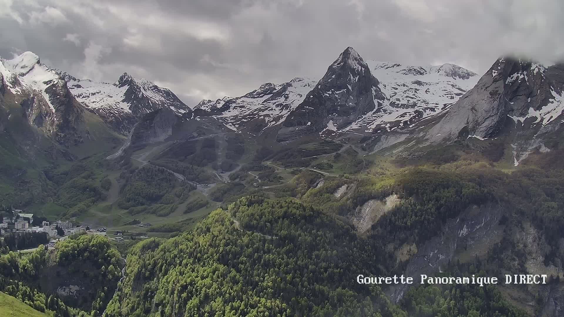 A valley filled with lush green forests and a small town sits nestled beneath a range of snow-capped mountains under a cloudy sky.