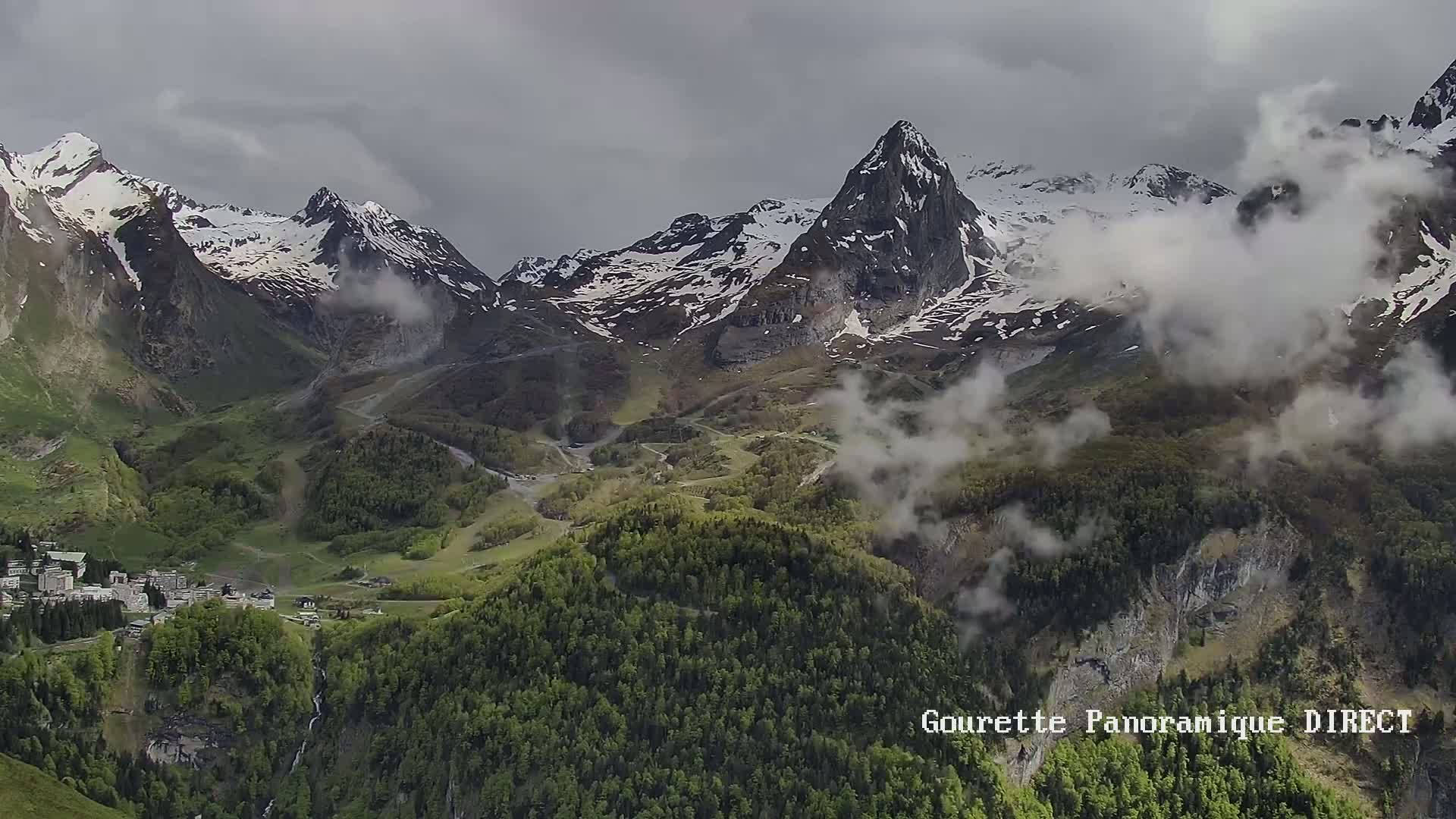 A mountain village nestled in a valley below snow-capped peaks partially obscured by clouds on an overcast day.