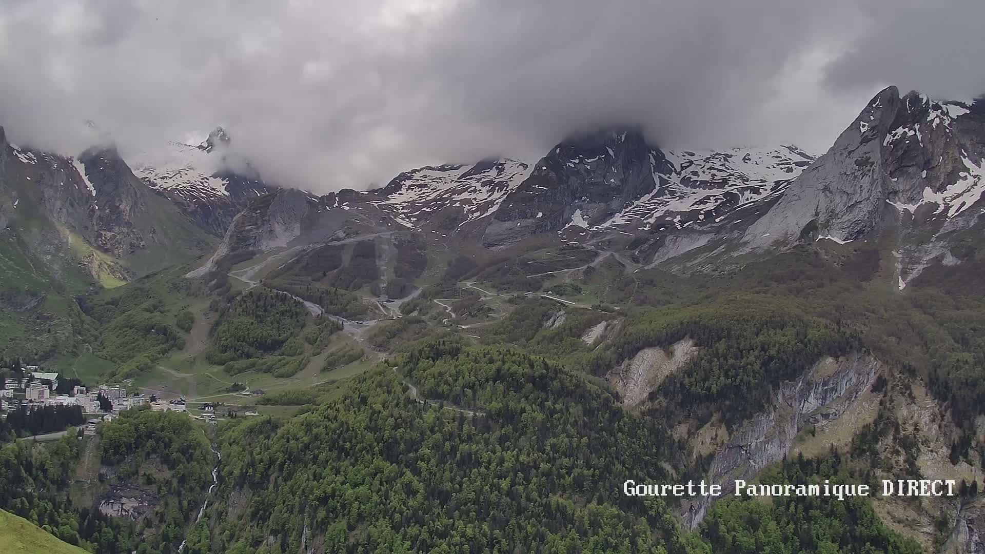 A mountain village nestled in a valley below snow-capped peaks partially obscured by clouds.