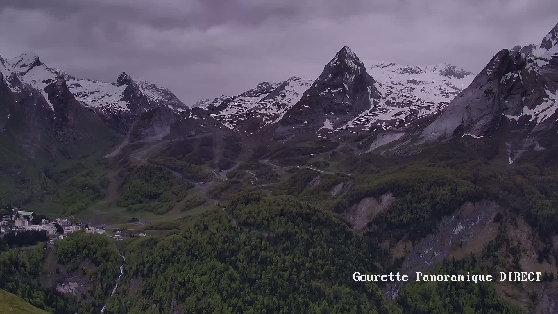 A mountain valley with snow-capped peaks under an overcast sky shows a small town nestled amongst lush green forests and waterfalls.