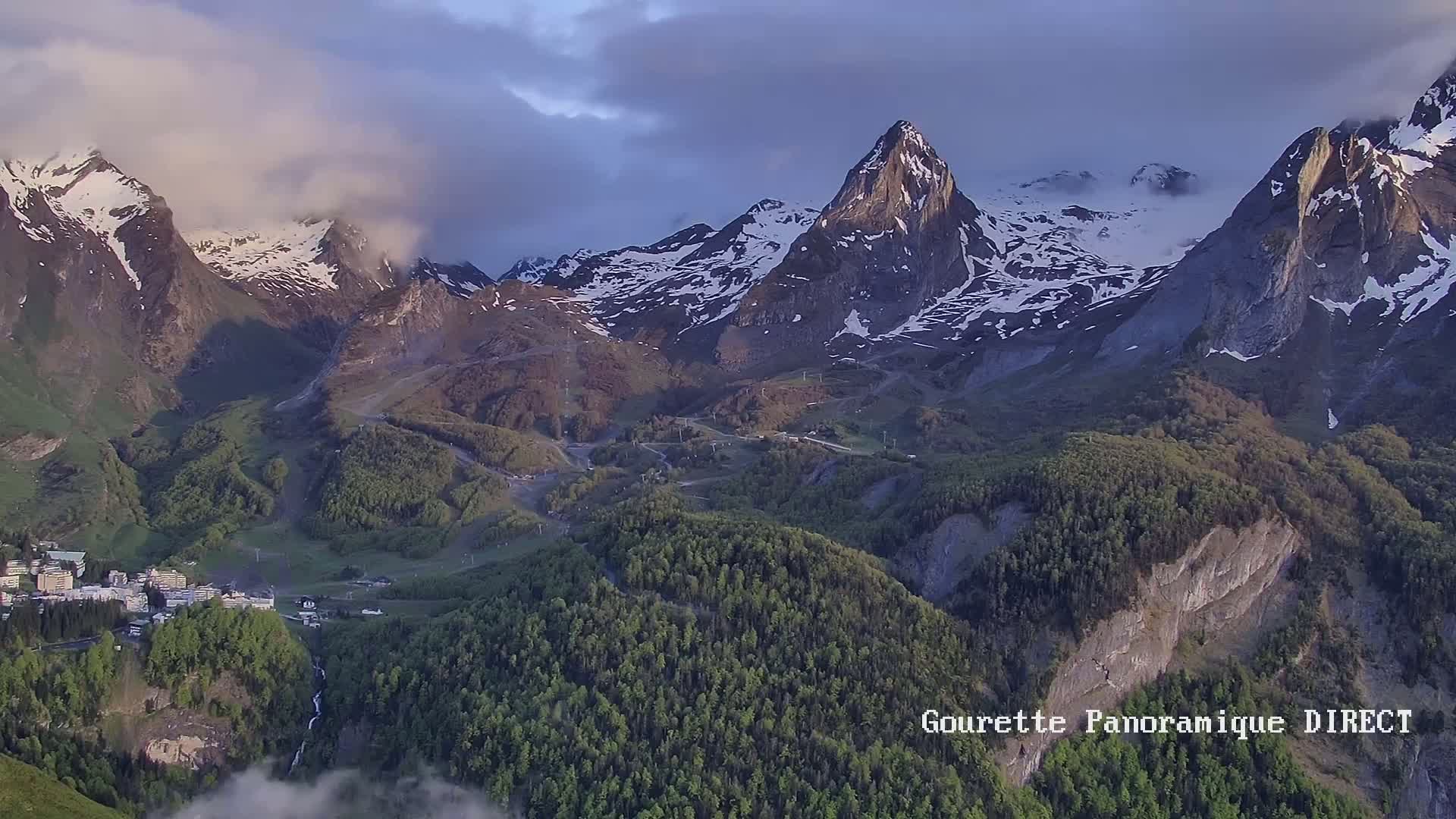 A mountain village nestled in a valley below snow-capped peaks, partially obscured by clouds under a partly cloudy sky.