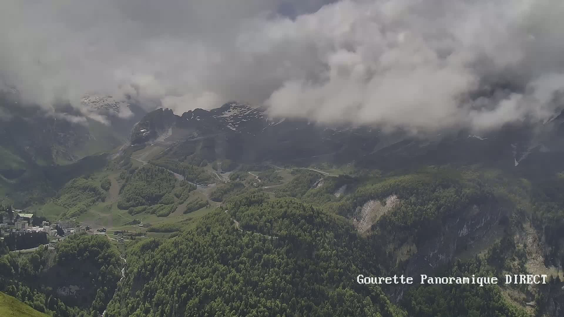 A mountainous landscape with green forests and a village nestled in a valley is partially obscured by low-hanging clouds.