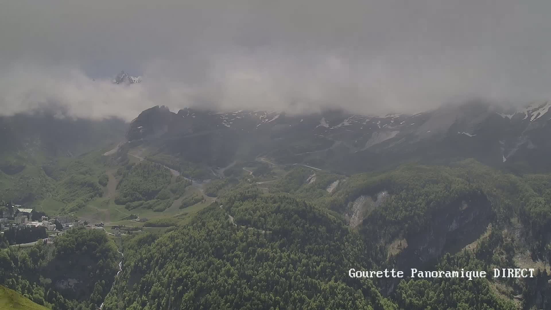 A misty, cloudy day reveals a mountainous landscape with a small town nestled in a valley below, surrounded by lush green forests and snow-capped peaks.