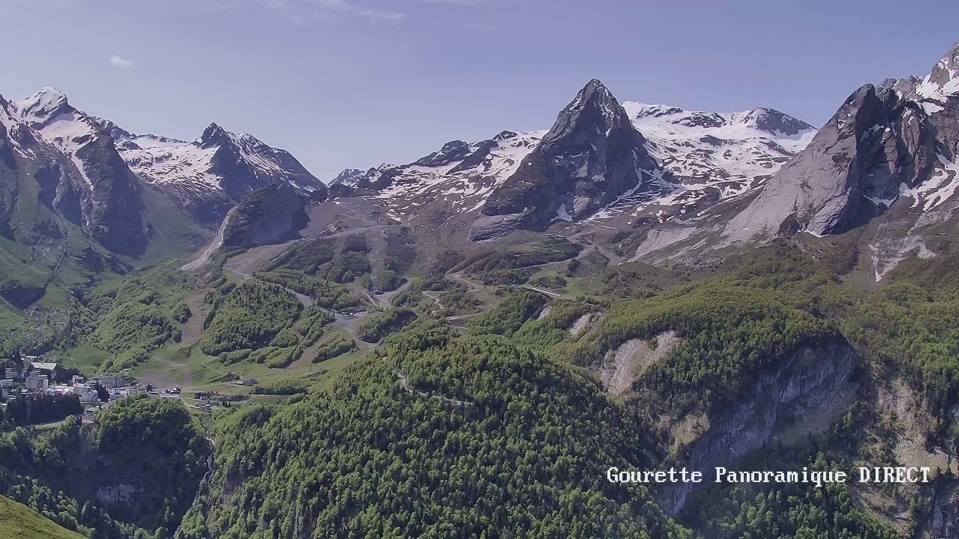 A mountain village nestled in a valley below snow-capped peaks on a sunny day.