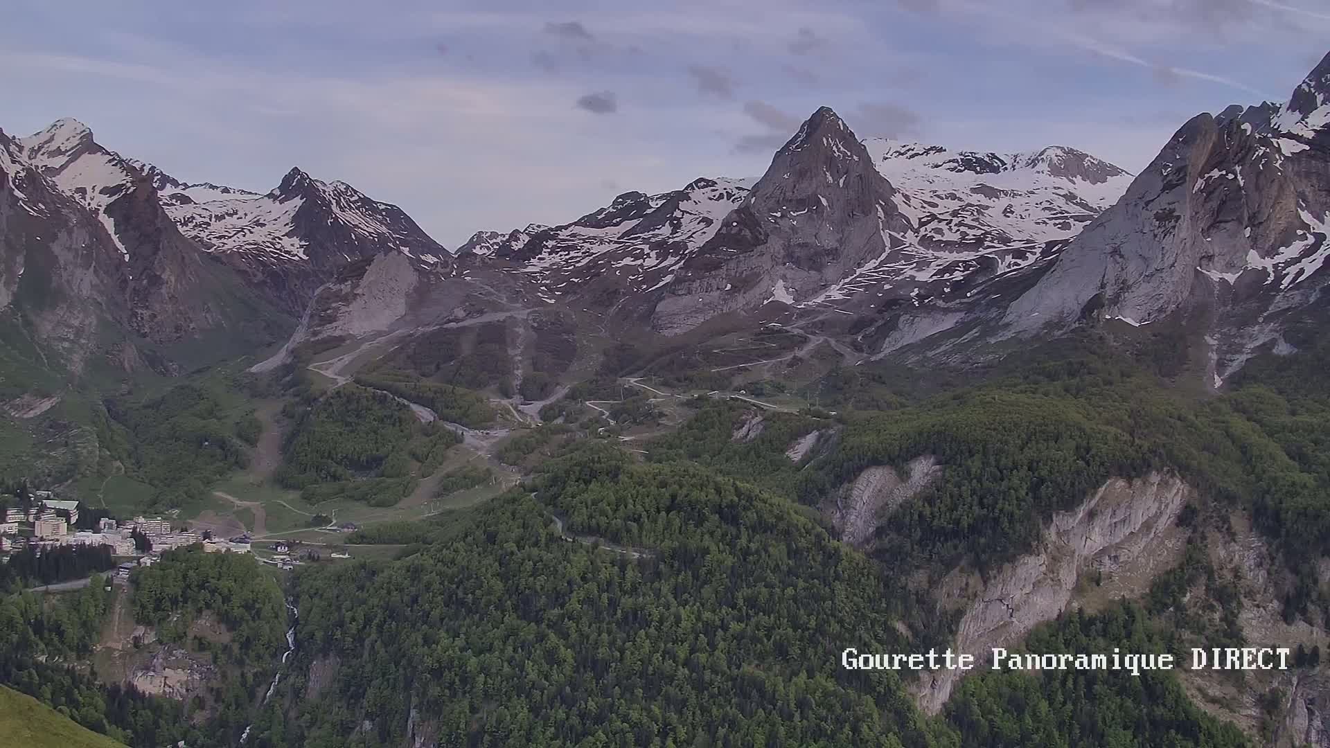 A mountain village nestled in a valley between snow-capped peaks under a partly cloudy sky.