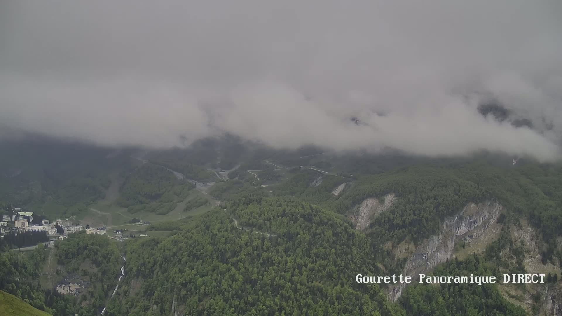 A foggy mountain landscape shows a valley with lush green forests, a small town, and winding roads.