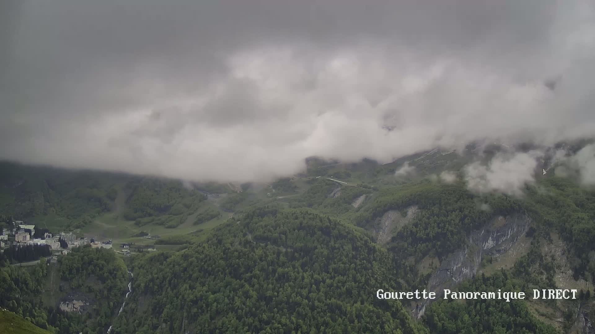 A mountain valley covered in a thick layer of fog and clouds, with a small village visible in the distance and lush green forests covering the slopes.