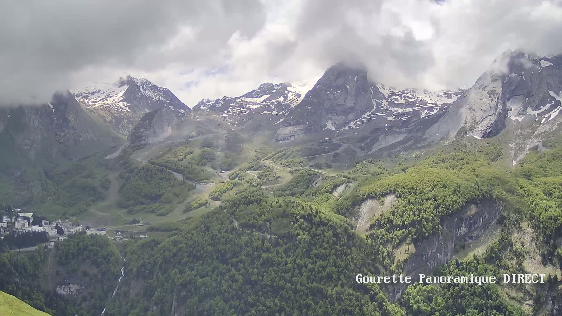 A partly cloudy day reveals snow-capped mountains and a valley covered in lush green vegetation, with a small town nestled at the valley's base.