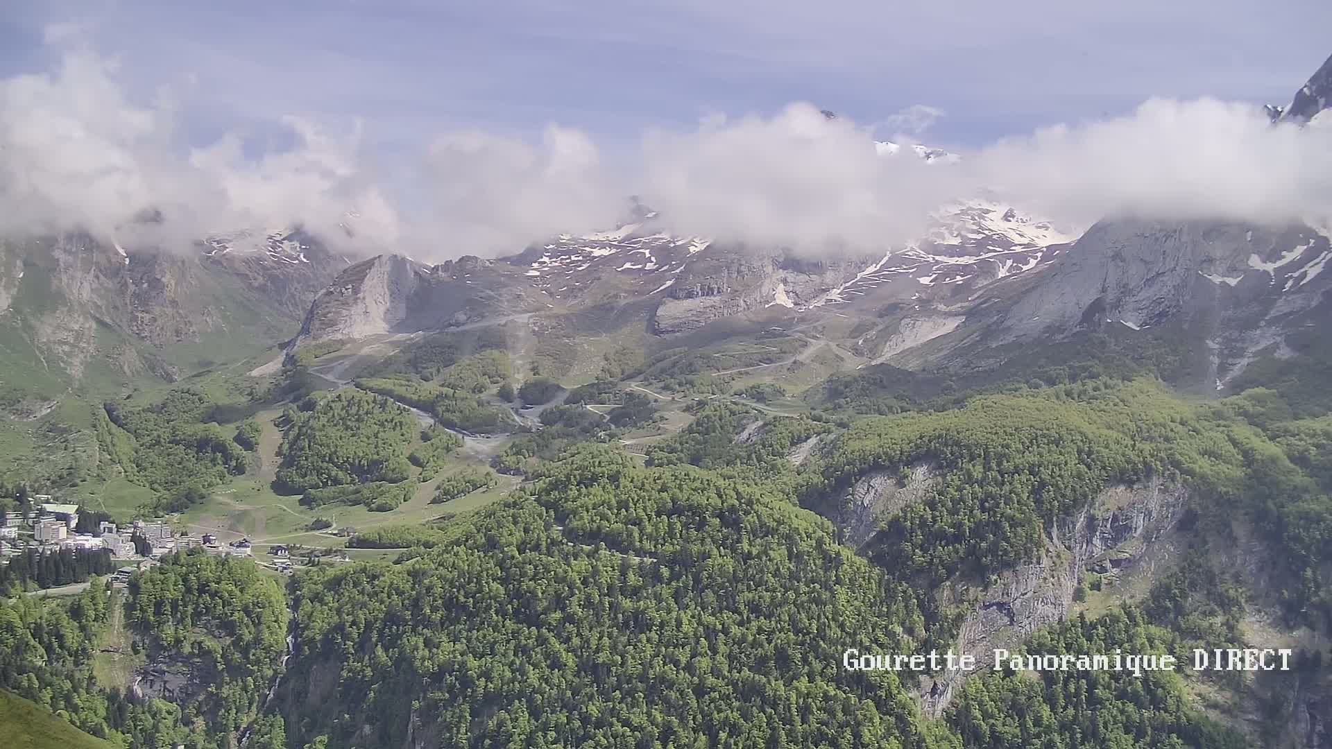 A mountain valley with a village nestled amongst lush green forests and snow-capped peaks partially obscured by clouds under a mostly clear sky.