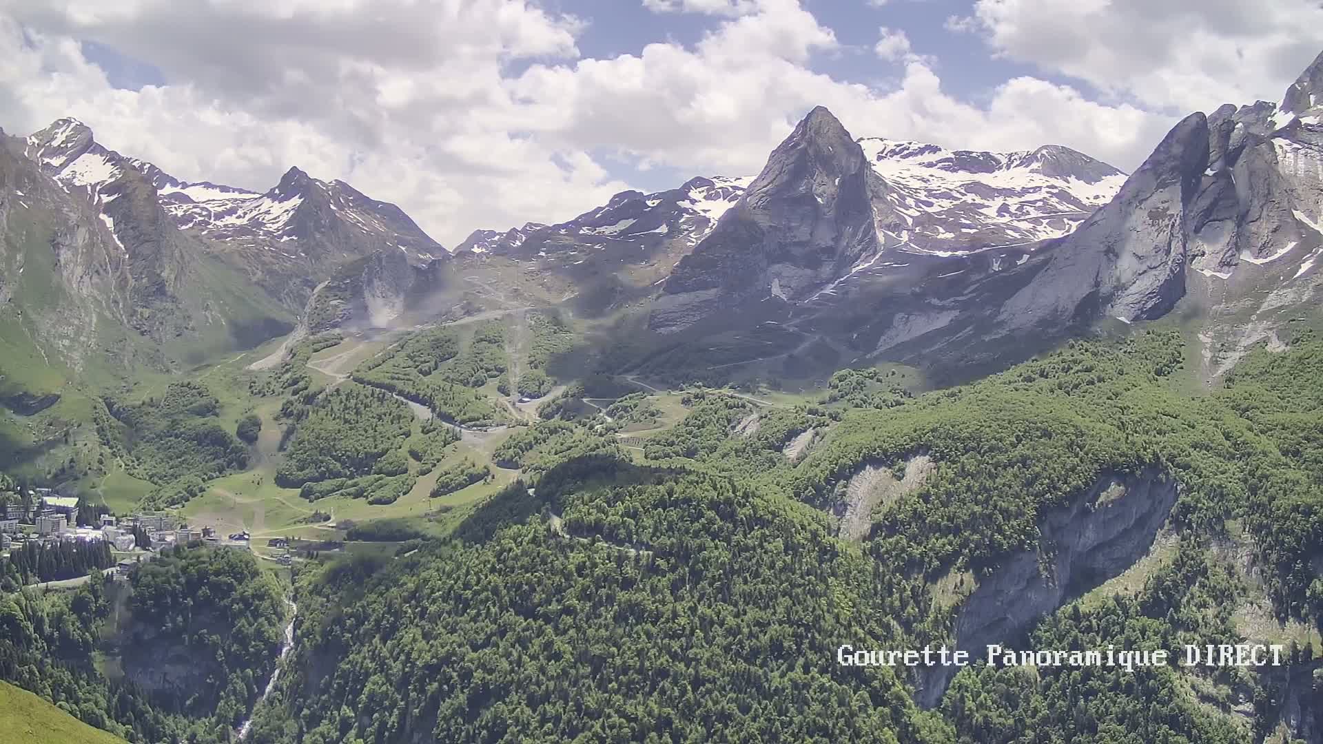 A partly cloudy day reveals a mountainous landscape with snow-capped peaks, lush green valleys, and a small village nestled within the mountains.