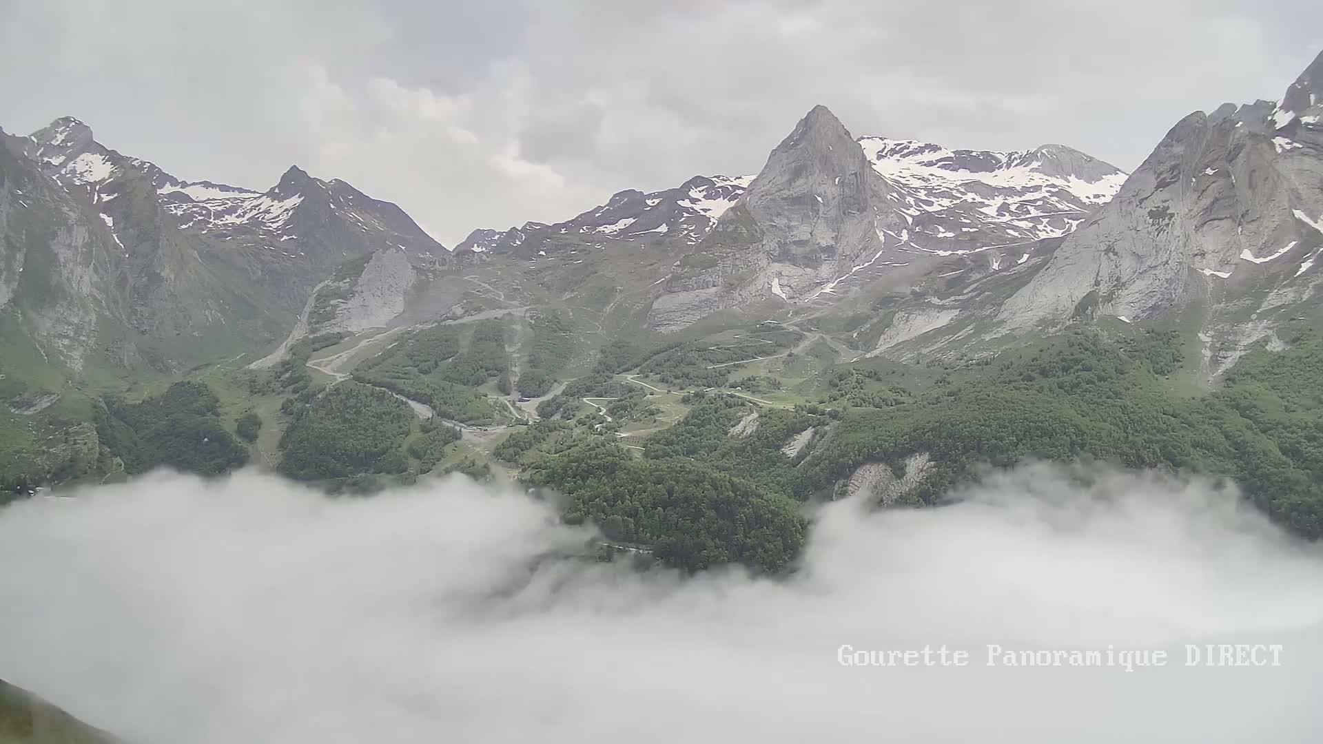 Snow-capped mountains and a valley covered in green vegetation are partially obscured by low-lying clouds under an overcast sky.