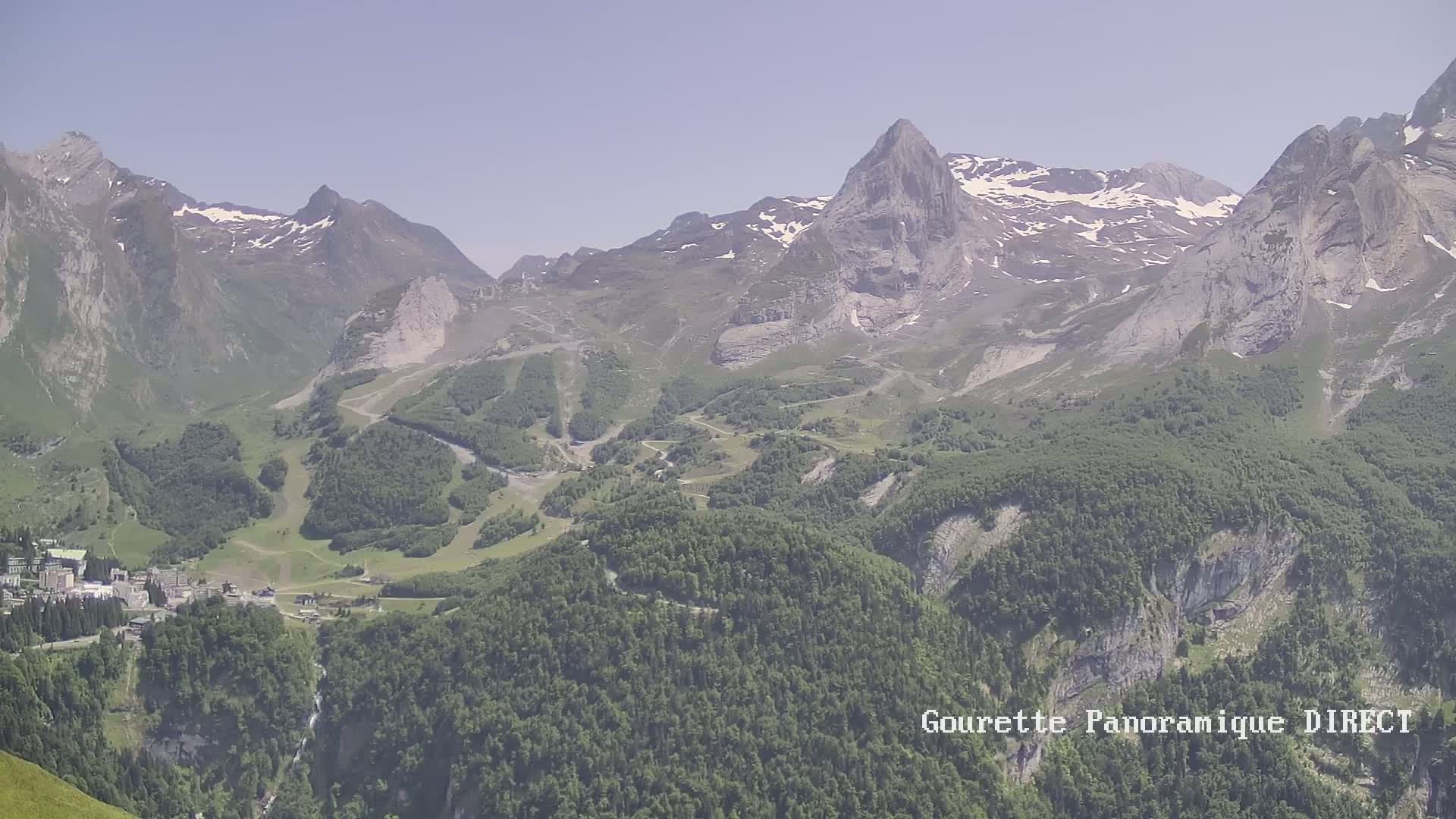 A mountain valley with snow-capped peaks, green forests, and a small village nestled within, under a clear sky.