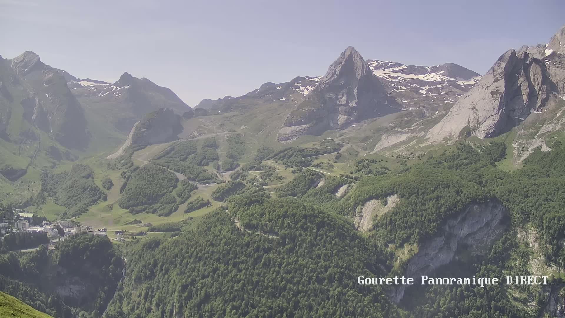 A hazy, sunny day reveals a mountainous landscape with a village nestled in a valley below snow-capped peaks and lush green forests.