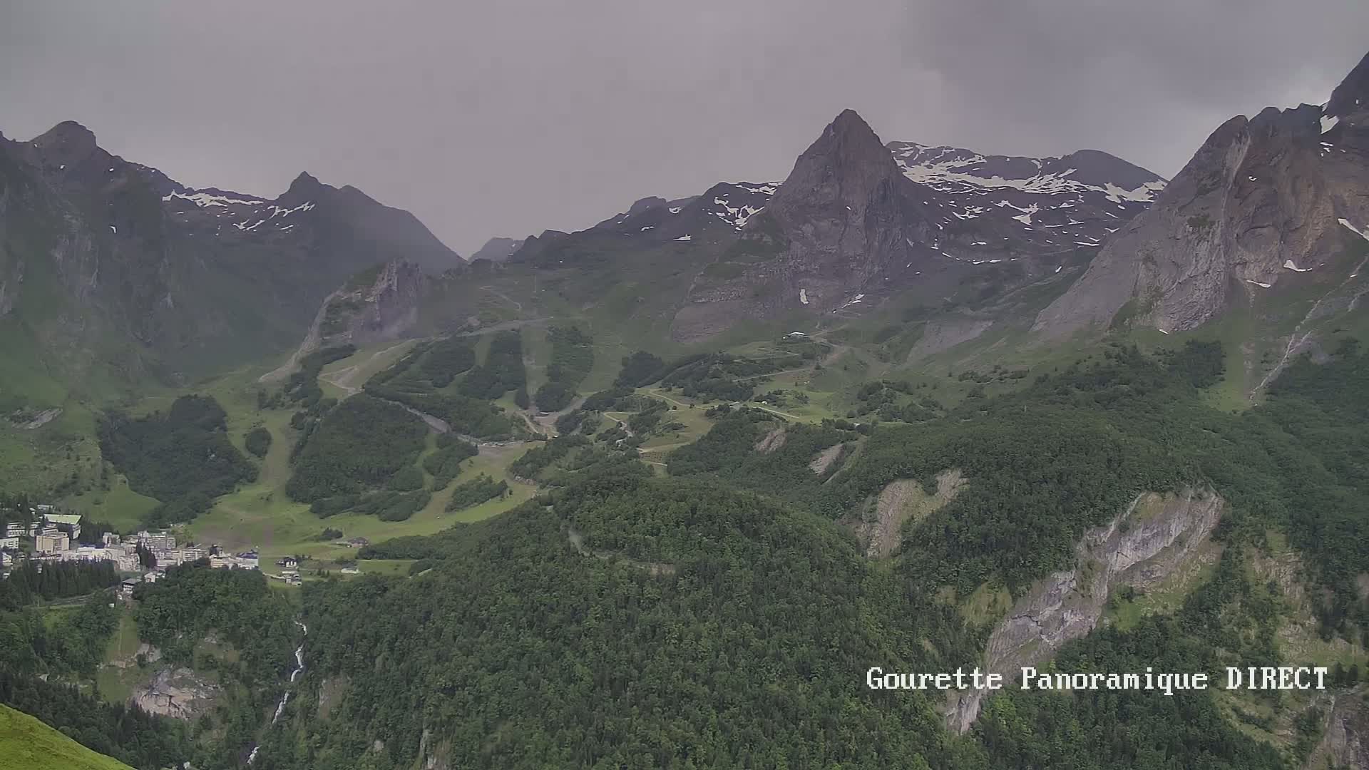 A mountainous landscape under an overcast sky shows a village nestled in a valley between steep, green slopes and rocky peaks with patches of snow.