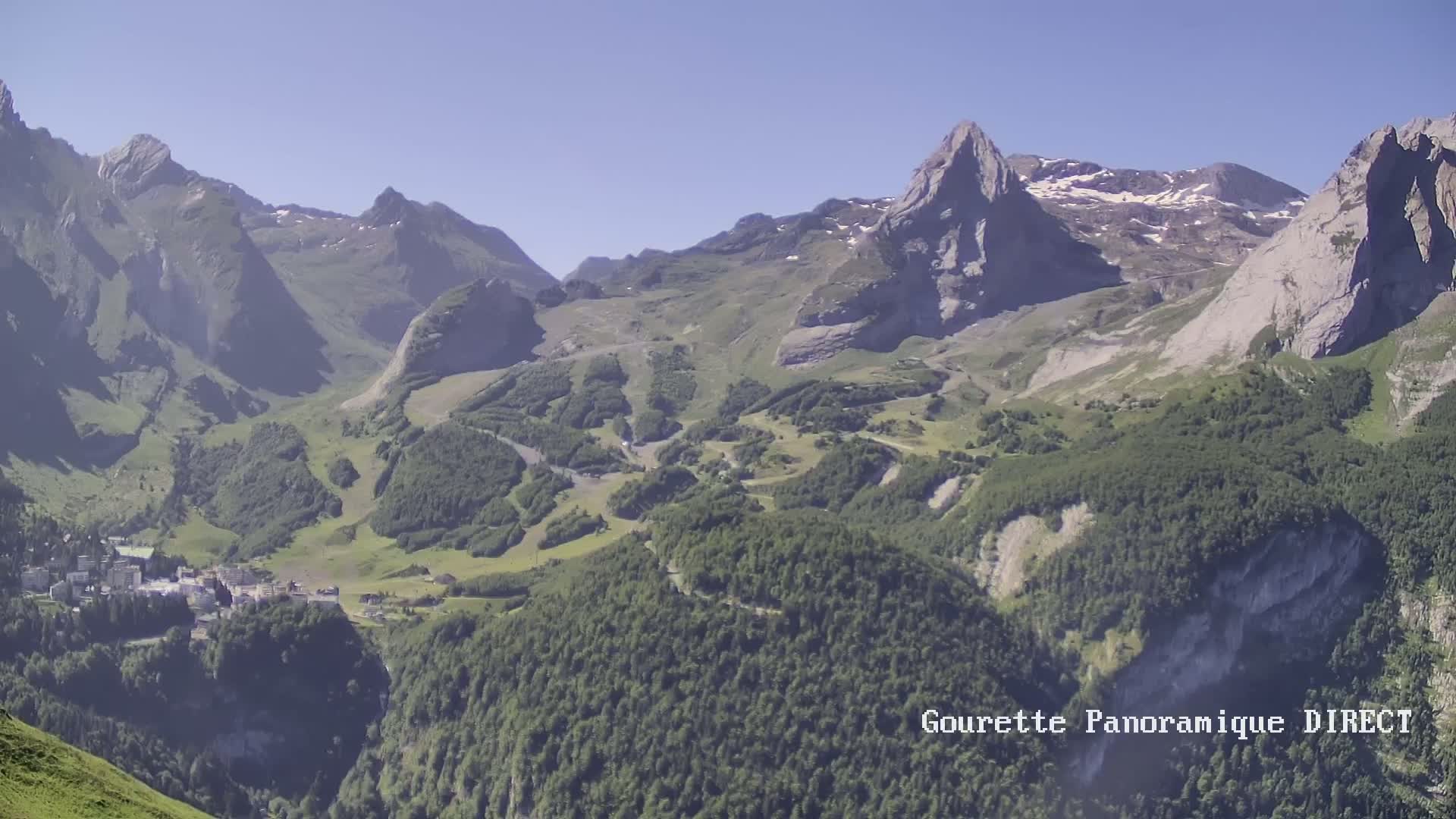 A high-angle view on a clear day shows a mountain valley with a small town nestled among green hills and rocky, snow-capped peaks.