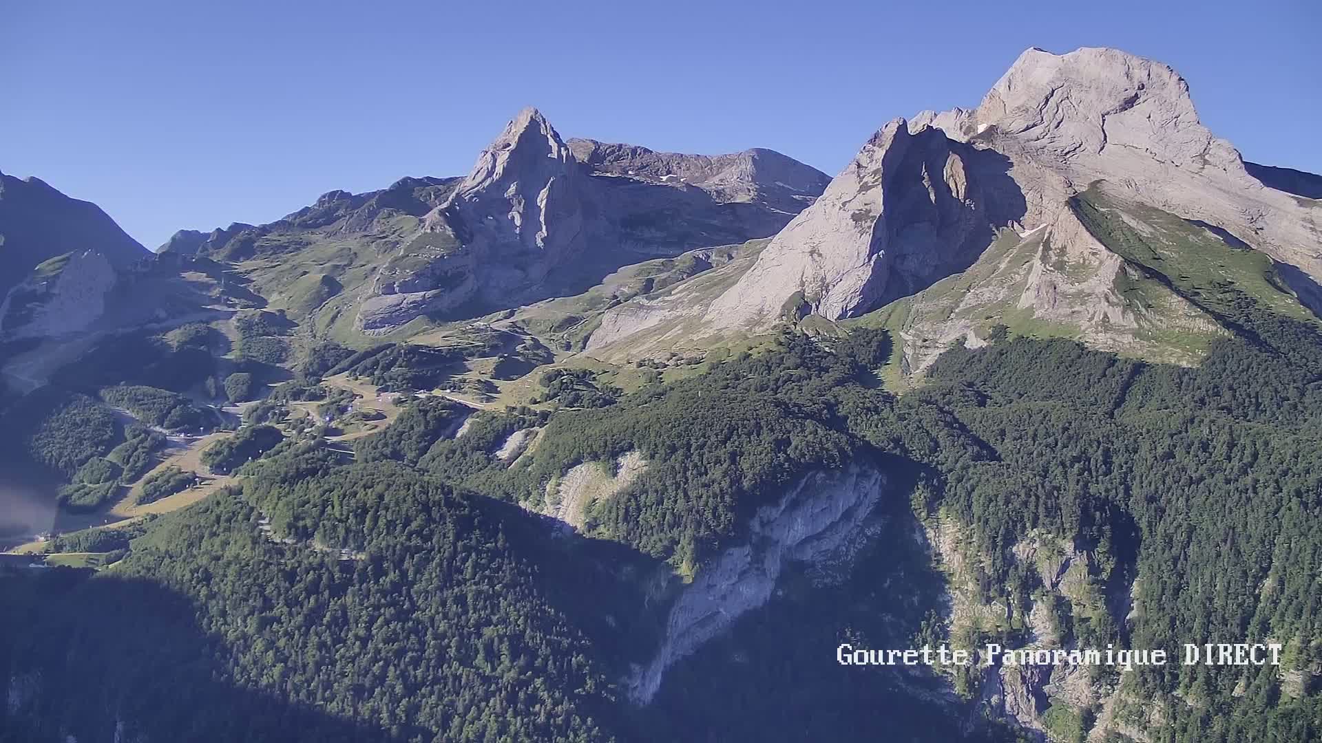 A high-angle view on a clear, sunny day shows a mountainous landscape with rocky peaks and verdant valleys covered in dense forest.