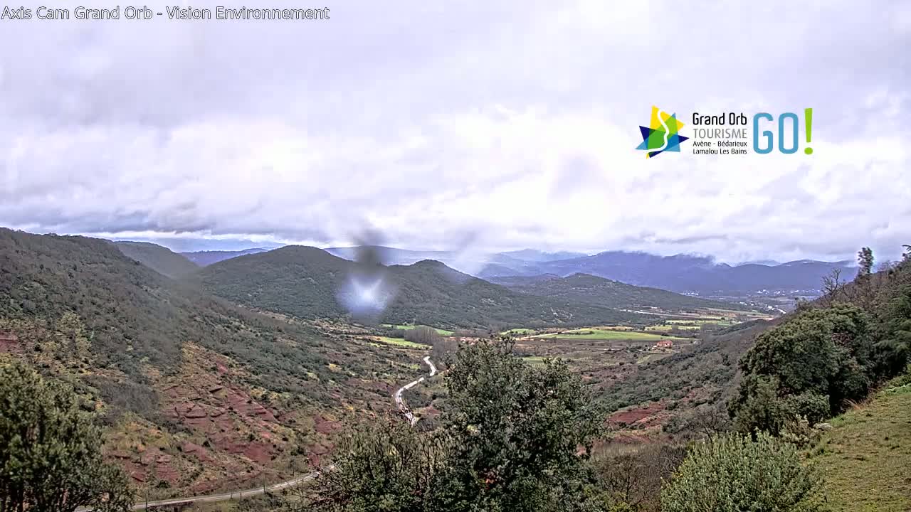 A winding road descends into a valley between rolling, tree-covered hills under a cloudy sky.