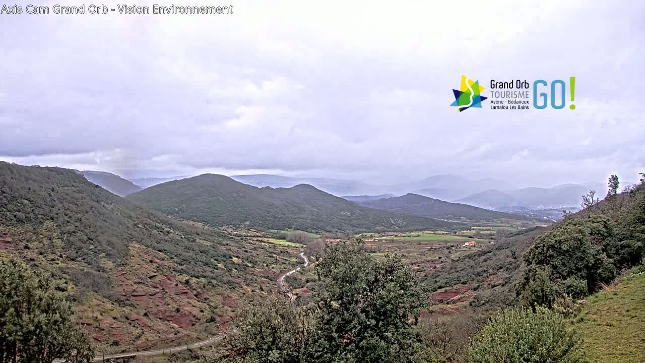 A winding road snakes through a valley of low, scrub-covered hills under a cloudy sky.