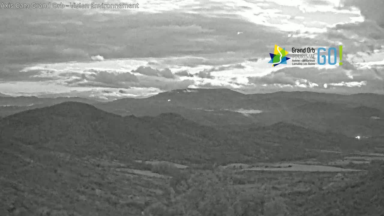 A grayscale image shows a mountainous landscape under a partly cloudy sky.