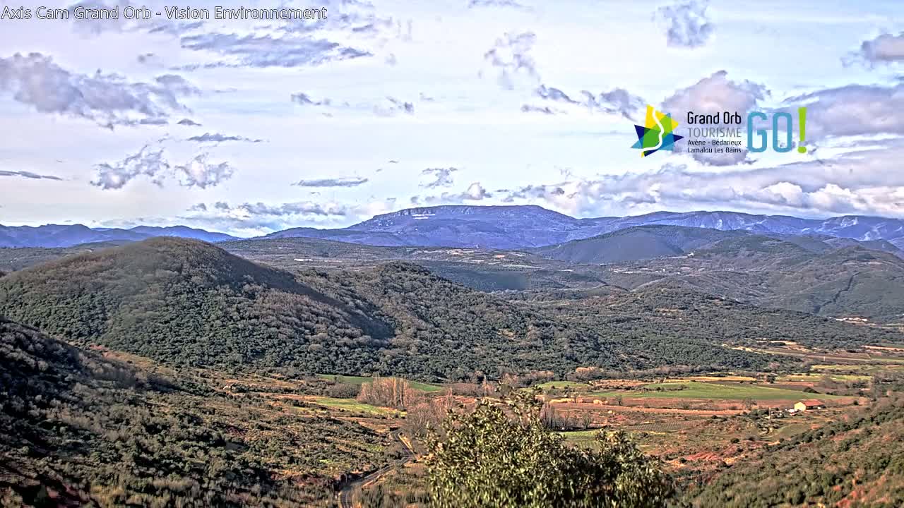 A partly cloudy day reveals a sweeping vista of rolling hills and distant mountains, covered in a mix of scrubby vegetation and cultivated fields.