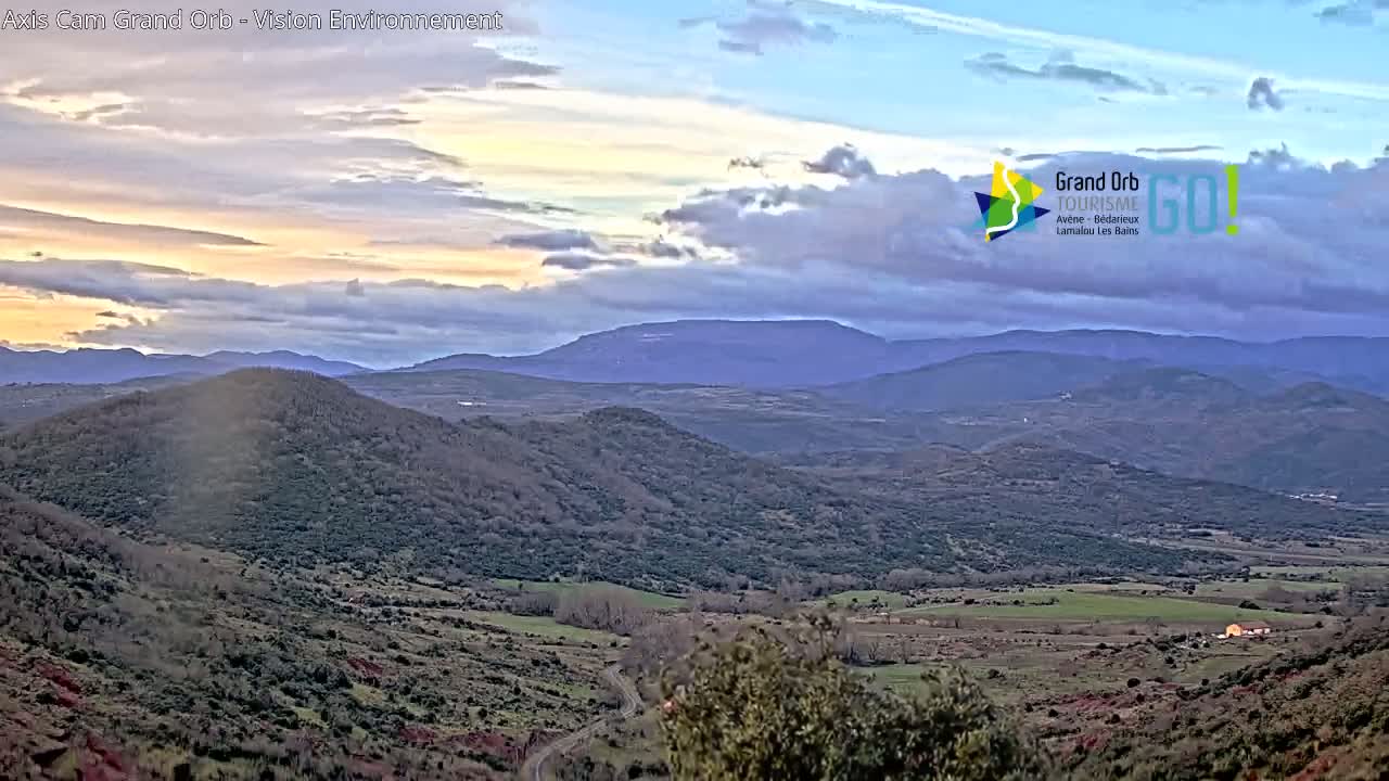 A panoramic view of rolling hills and distant mountains under a partly cloudy sky with a soft, pale sunset.