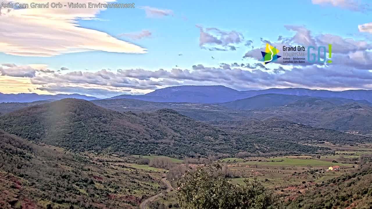A partly cloudy sky overlooks a vast, hilly landscape of muted greens and browns, with distant mountains in the background.