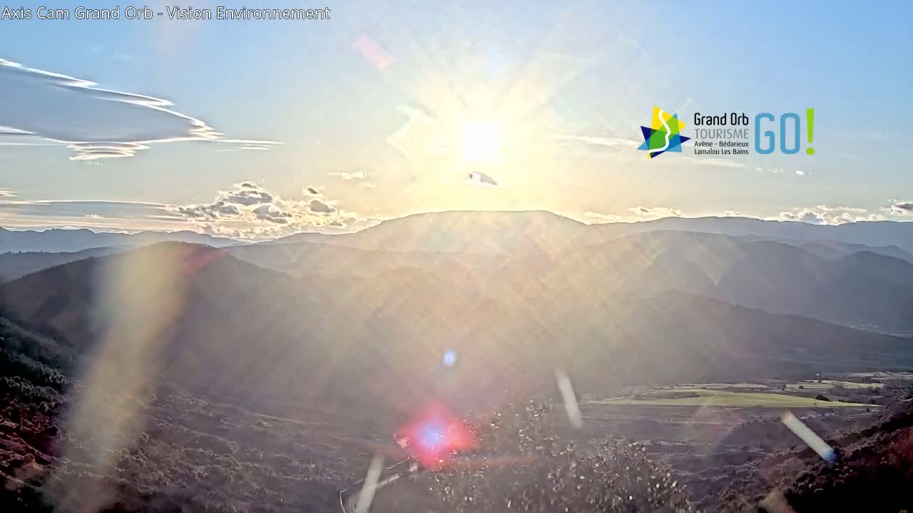 A bright sun shines over a mountainous landscape at sunset, with some thin clouds in the sky.
