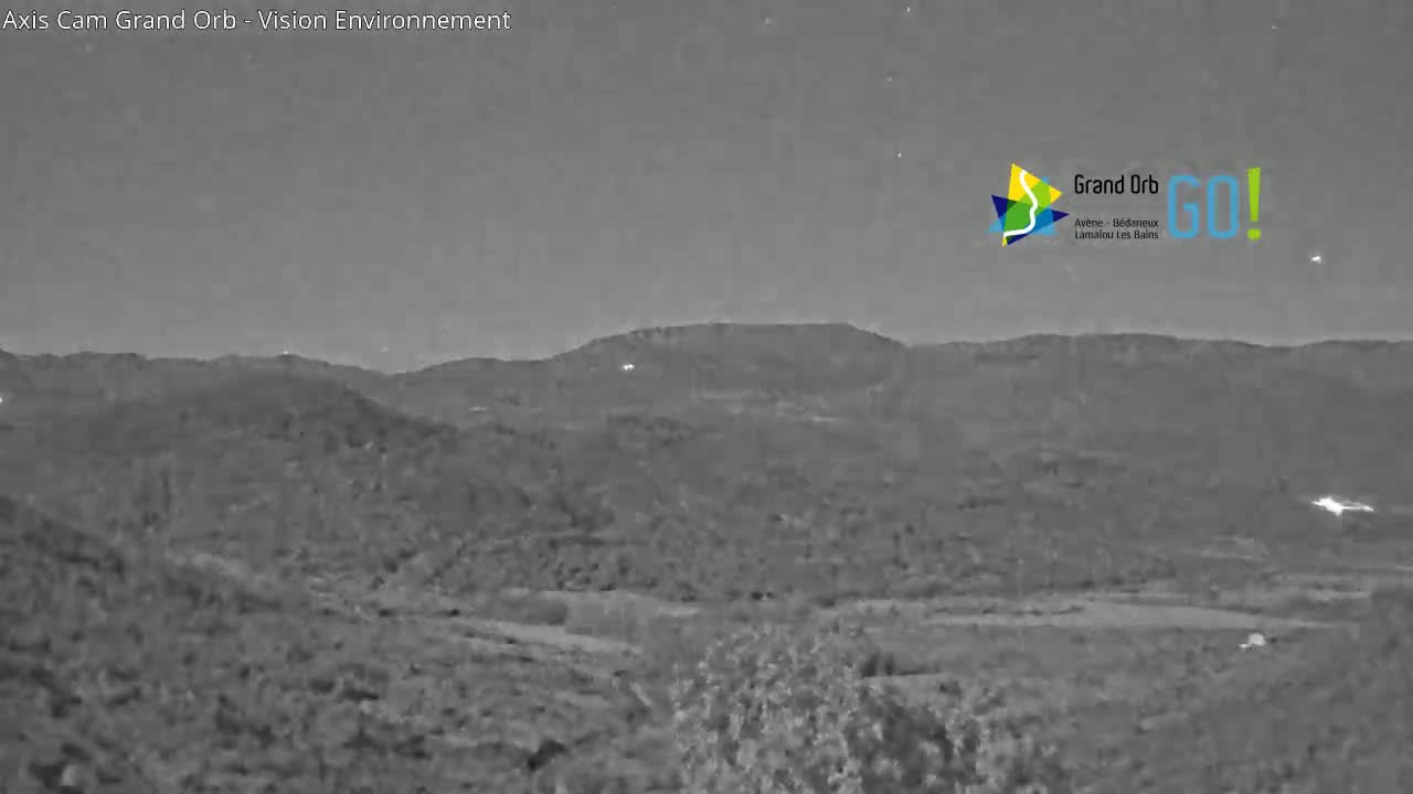 A nighttime, grayscale view of a hilly landscape under a mostly clear sky.