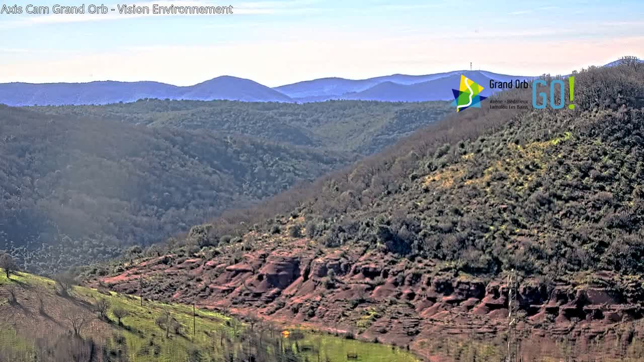 A view of a hilly landscape under a mostly clear sky shows reddish-brown rock formations and sparsely wooded hillsides, with distant mountains in the background.