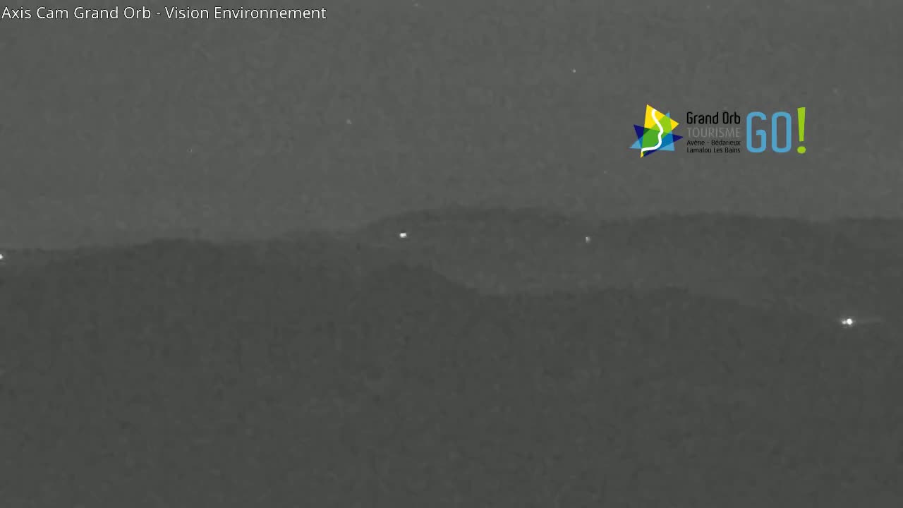 A nighttime, low-light image shows a dark, hilly landscape with a few small, distant lights visible.