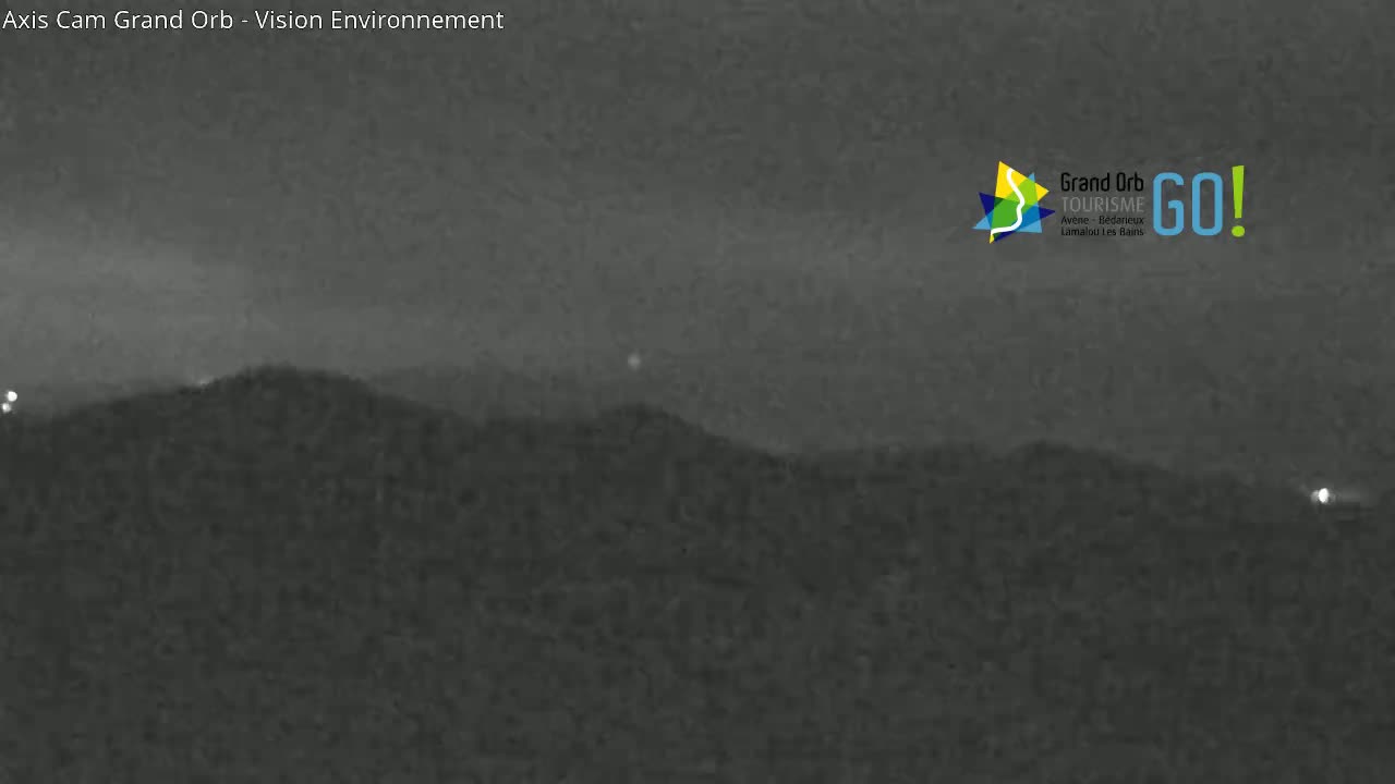 A nighttime, low-light image shows dark hills or mountains under a hazy, dark sky with a few distant, faint lights visible.