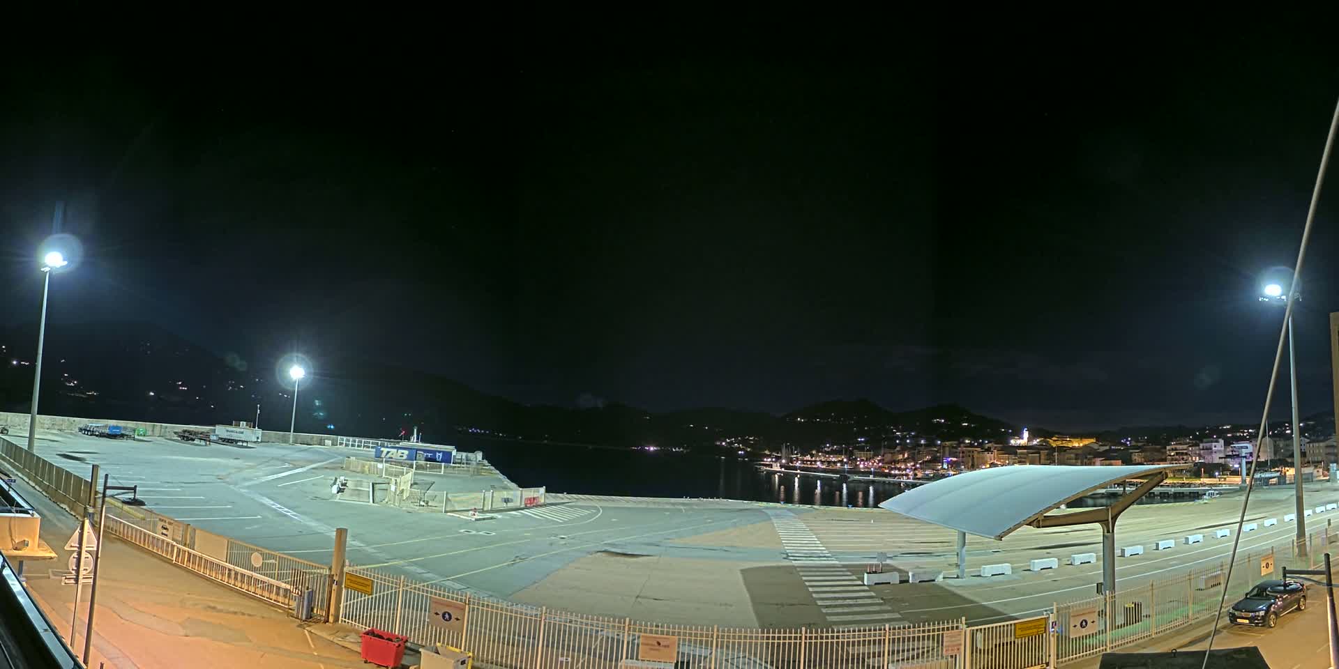 A nighttime, brightly lit, empty port area with a few vehicles, overlooks a dark body of water and a distant city illuminated by lights under a clear, dark sky.