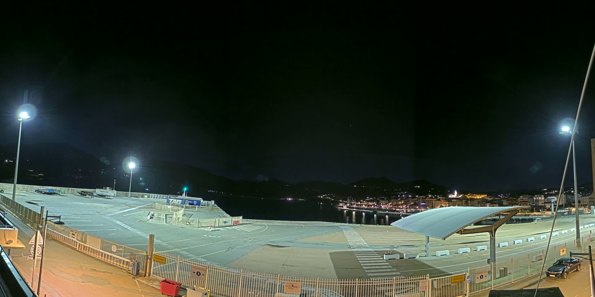 A nighttime, brightly lit, empty port area with a few vehicles and a covered waiting area overlooks a calm body of water and a town nestled against hills in the distance.