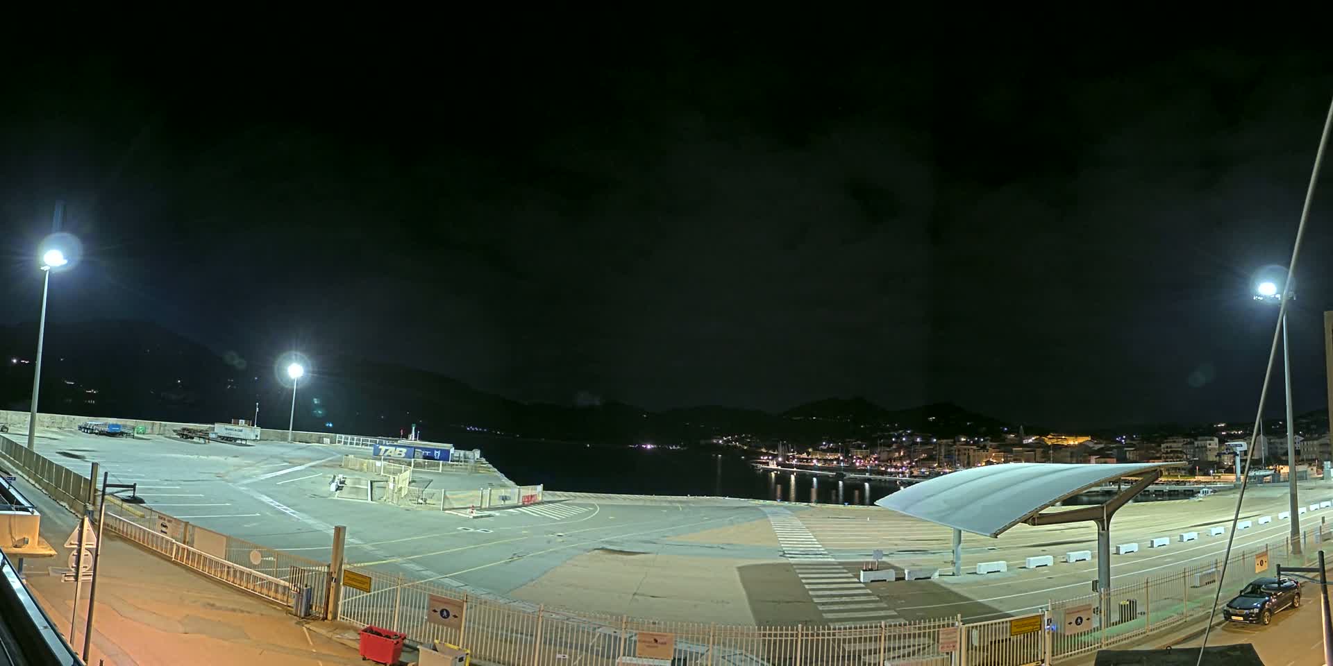 An empty, brightly lit port area at night, overlooking a dark body of water and a distant town under a partly cloudy sky.