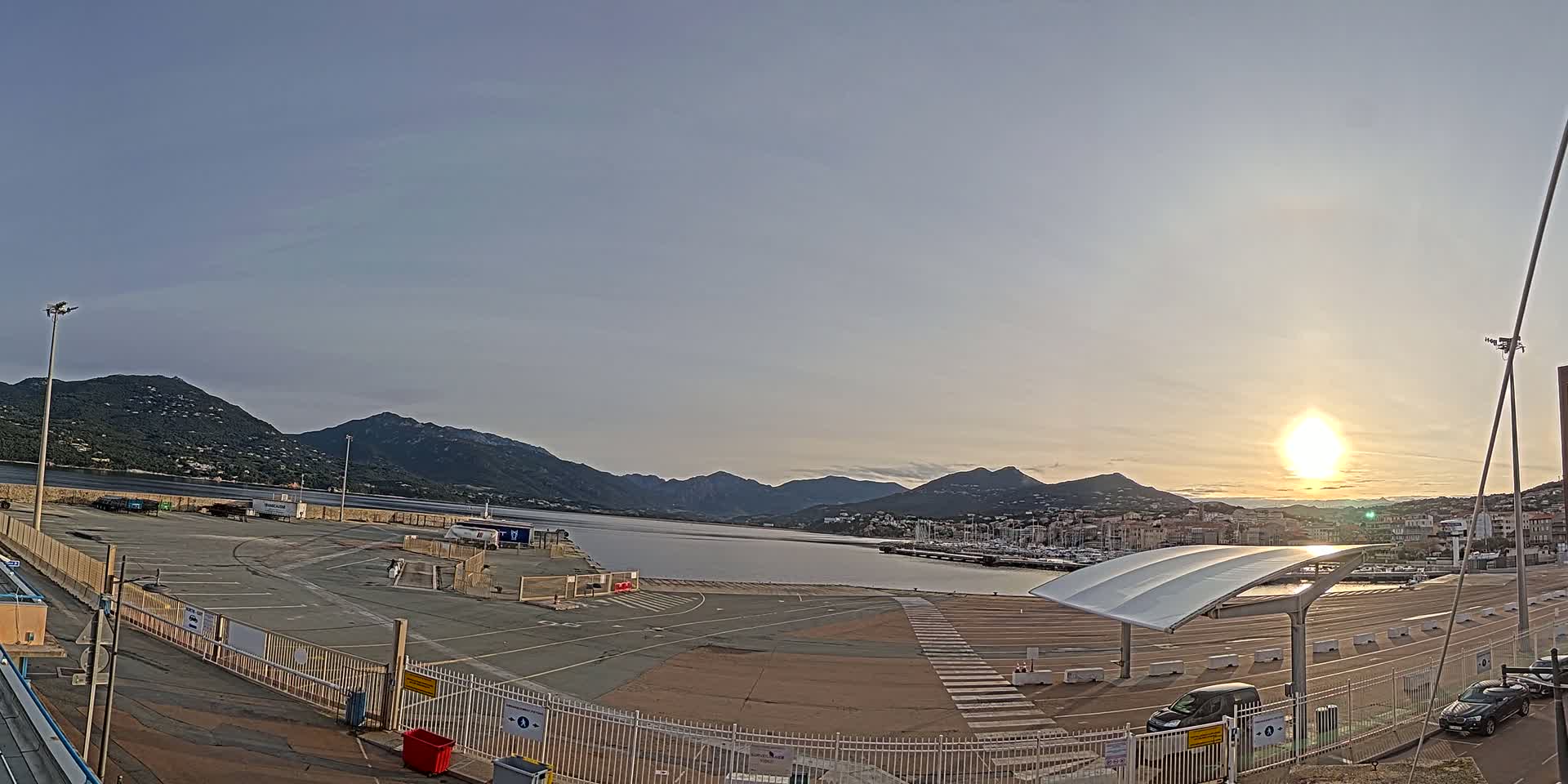 An empty port area with a modern canopy structure overlooks a calm body of water surrounded by mountains under a clear, sunrise sky.