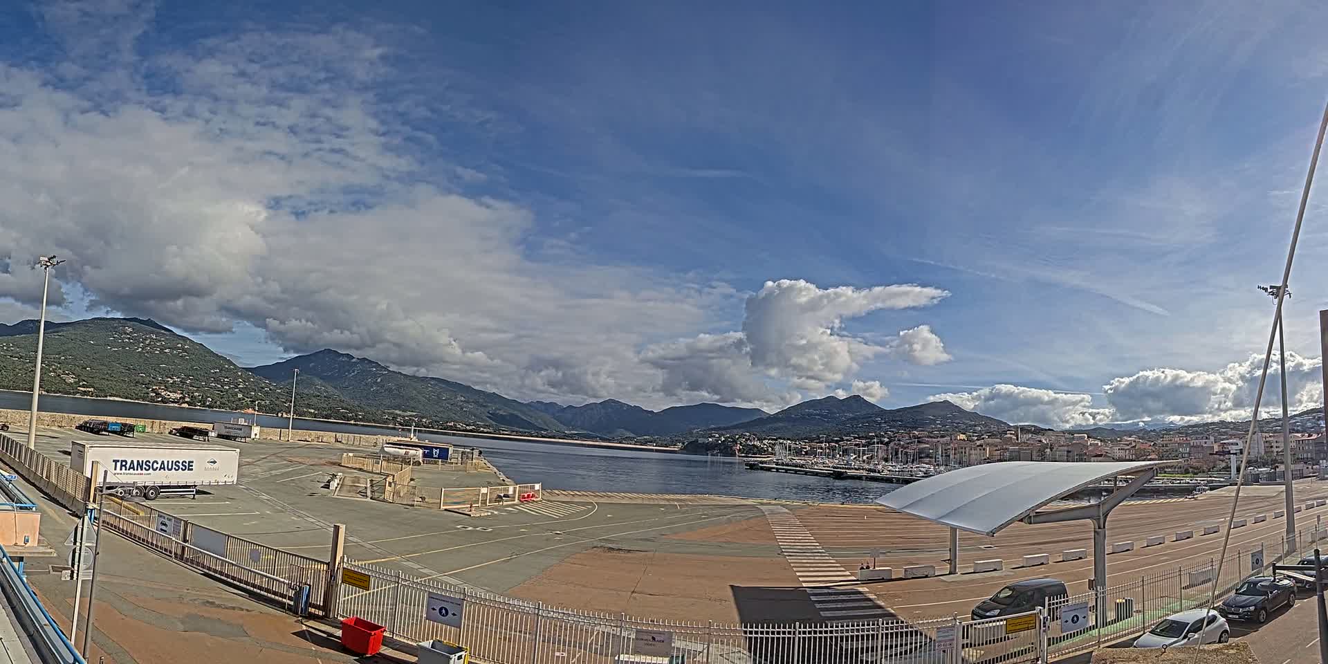 A mostly sunny day with puffy clouds over a harbor, mountains, and a large paved area with several vehicles parked.