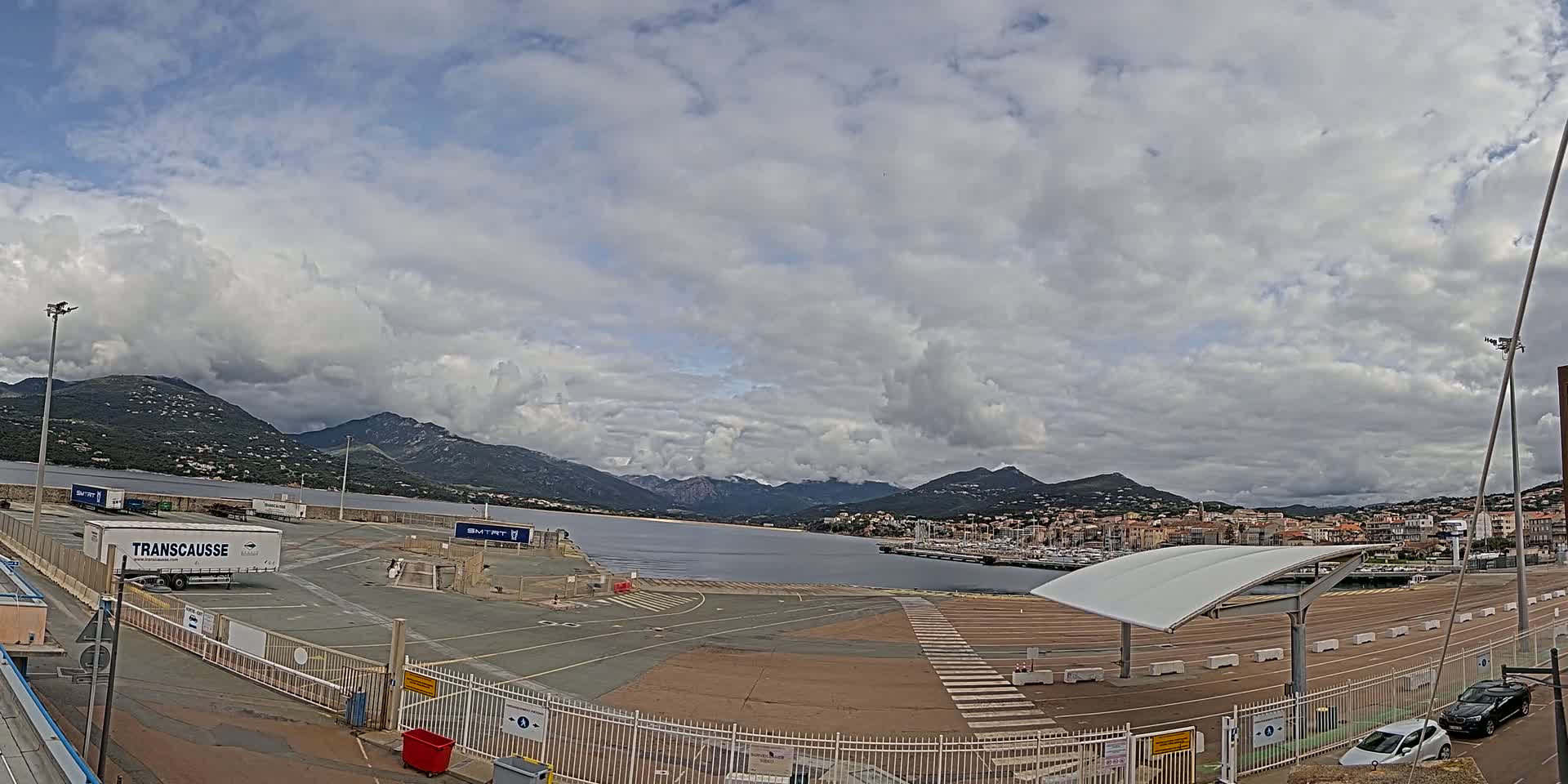 A mostly cloudy day overlooks a port area with a few vehicles and a large body of water surrounded by mountains.