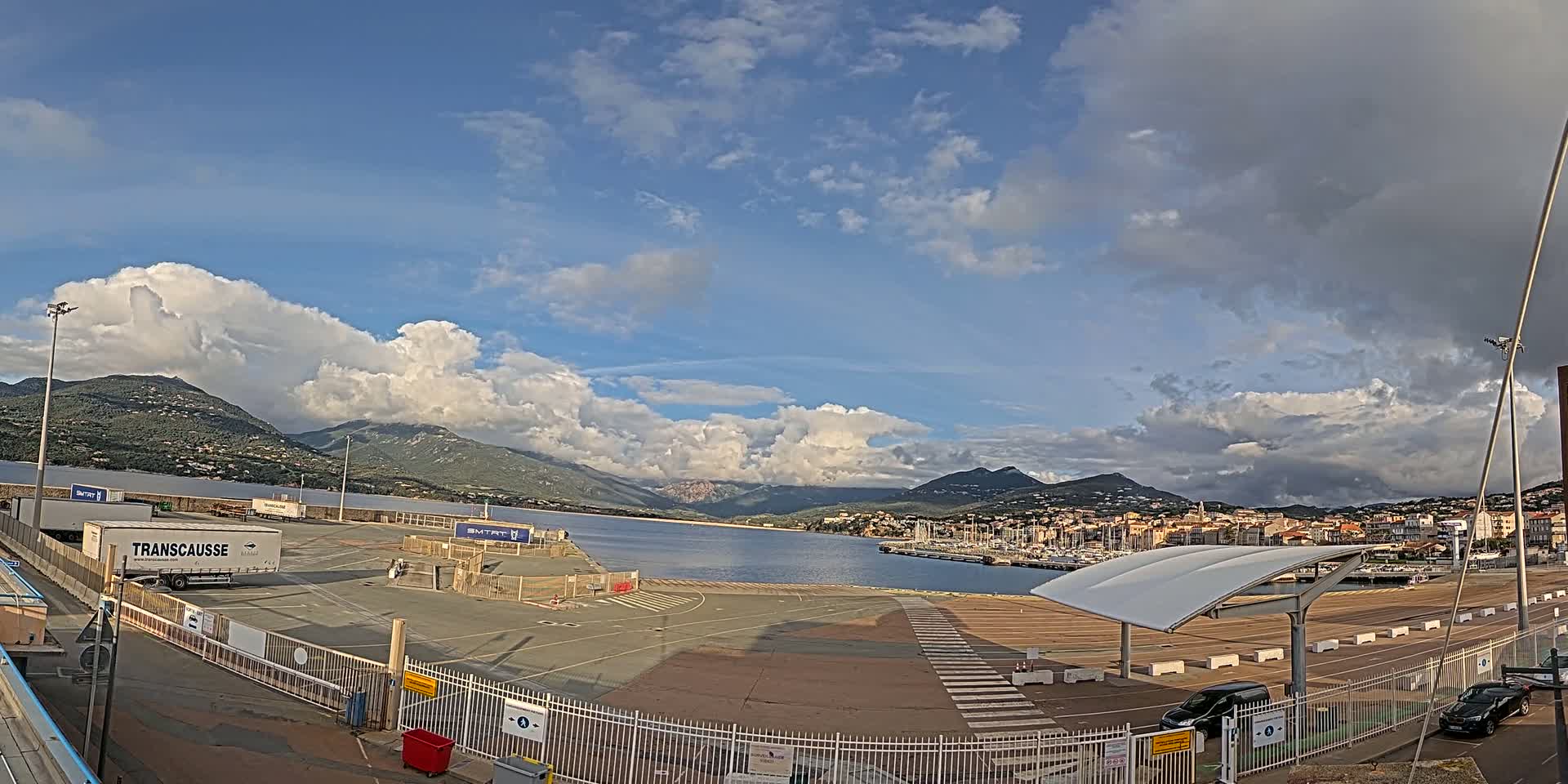 A mostly sunny day with puffy clouds over a harbor in a town nestled beside mountains, featuring a large, empty parking lot with several trailers and cars.