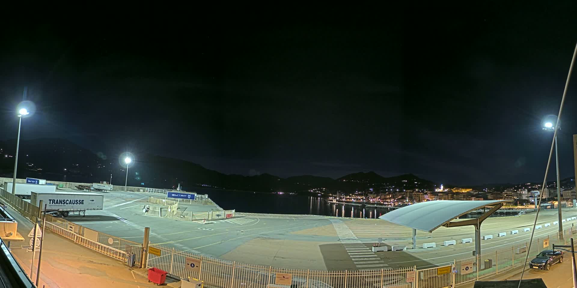 A nighttime view of an empty port area with several trailers, a covered waiting area, and a town across the water under a dark, clear sky.