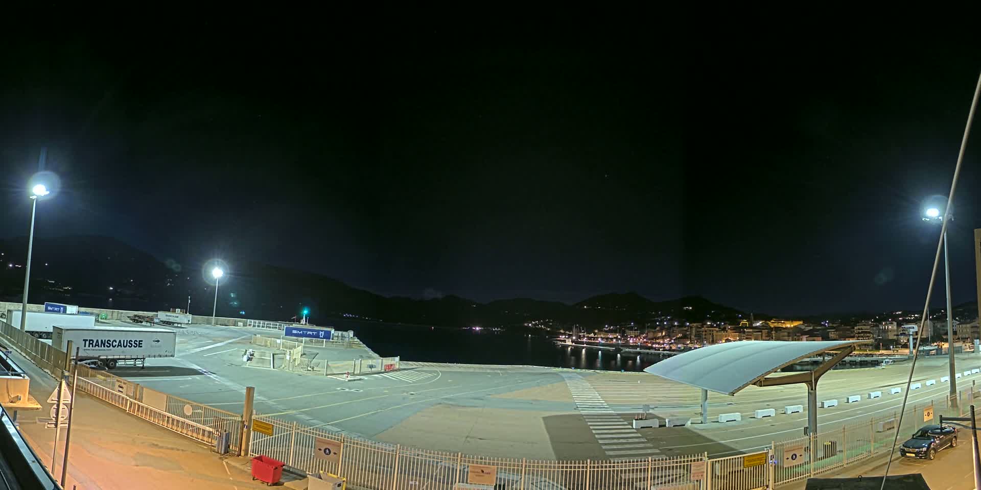 An empty, brightly lit ferry terminal at night overlooks a calm body of water and a town nestled against dark hills under a clear, starlit sky.