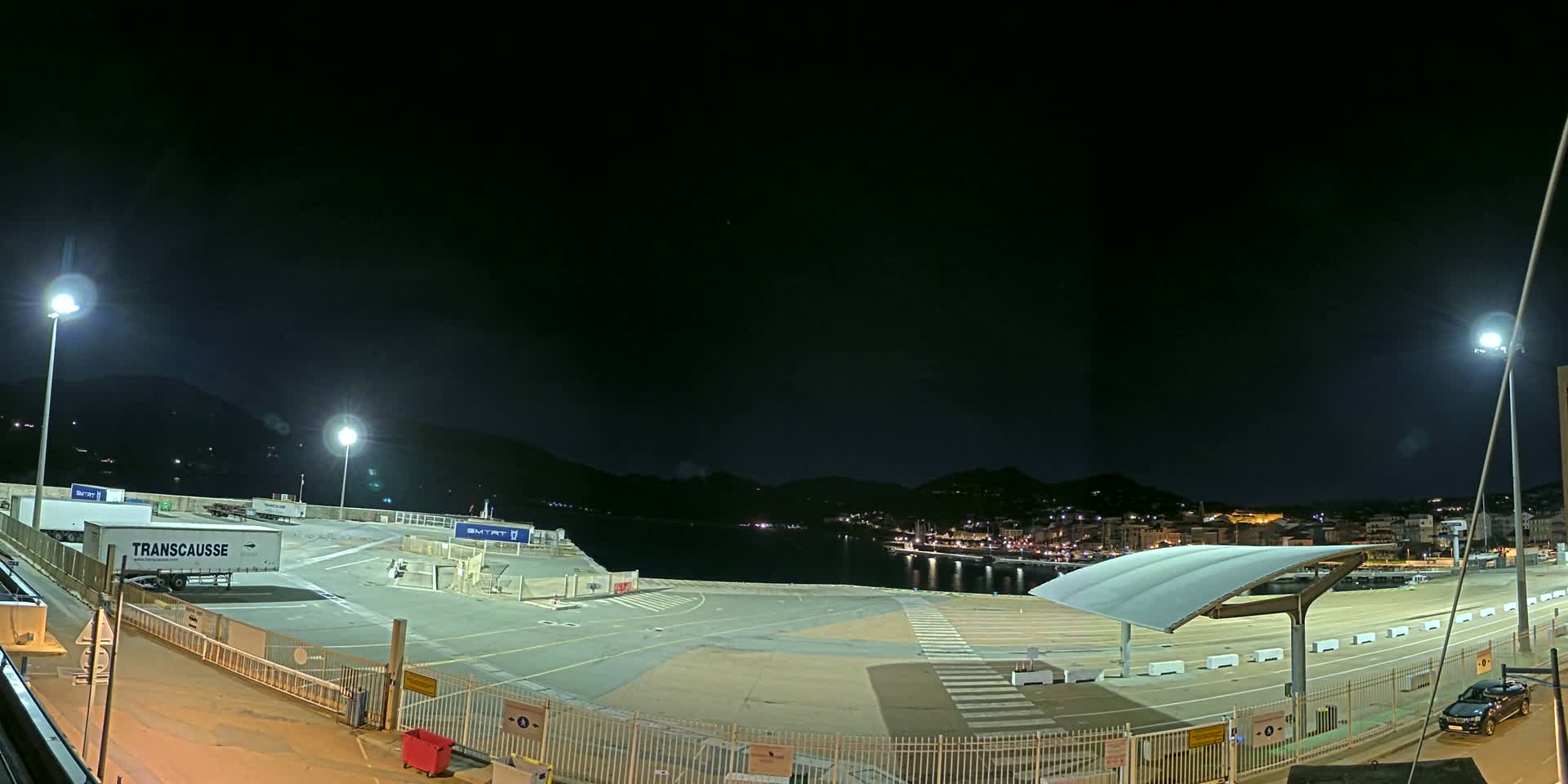 A nighttime view of a port area with several trucks, a covered waiting area, and a town illuminated across the water under a clear, dark sky.