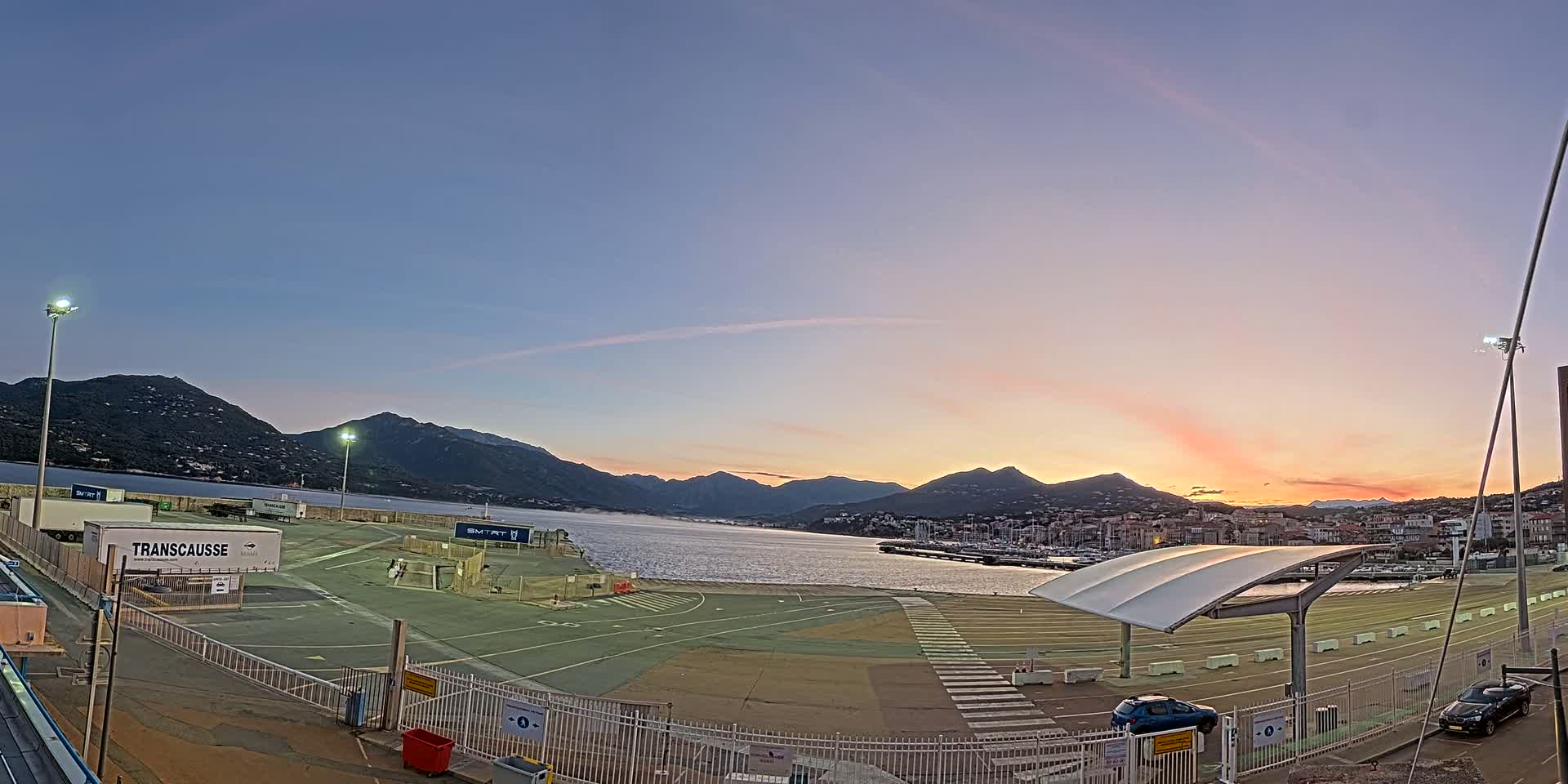 An empty ferry terminal at sunrise, overlooking a calm body of water and mountains in the distance.