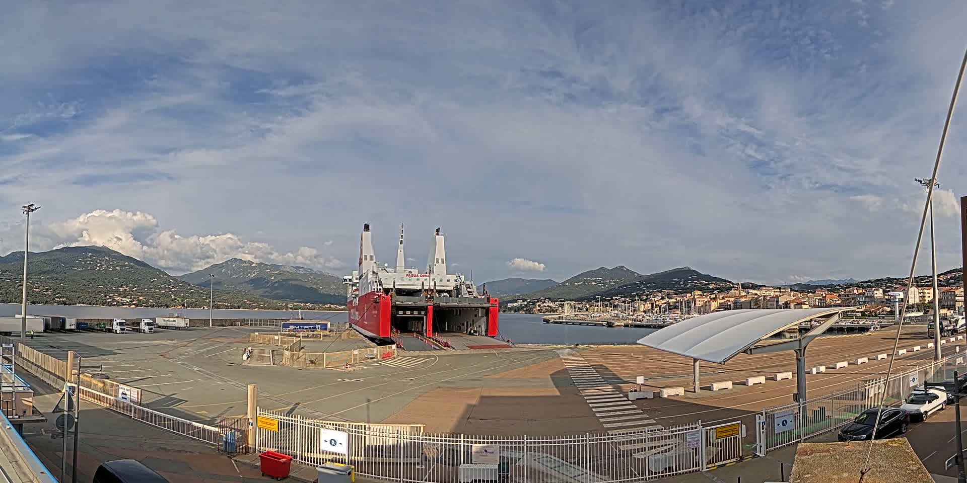 A large red and white ferry is docked at a port, with mountains in the background under a partly cloudy sky.