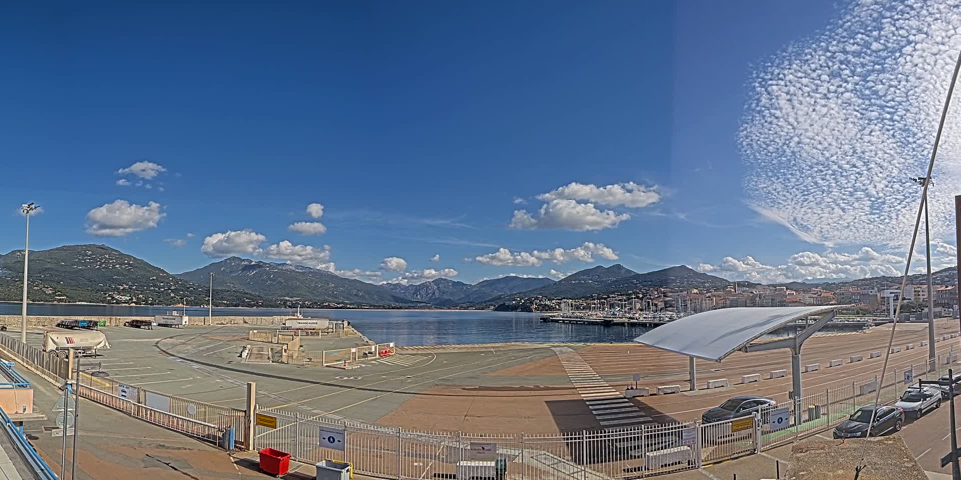 A mostly sunny day shows a harbor with a large empty paved area, some parked cars, and mountains in the distance.
