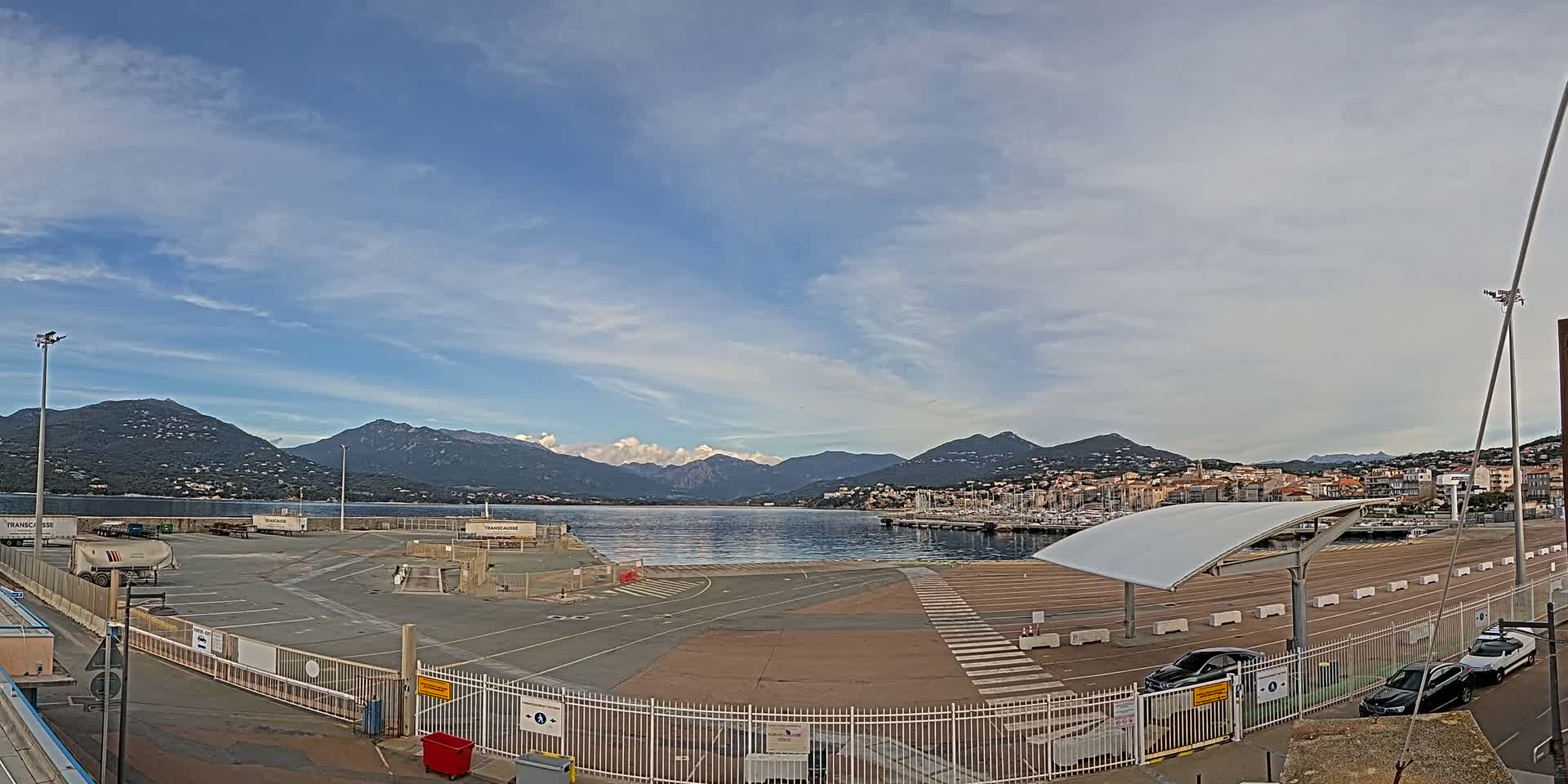 A mostly empty paved port area with a few cars, enclosed by a fence, overlooks a calm body of water and mountains under a partly cloudy sky.