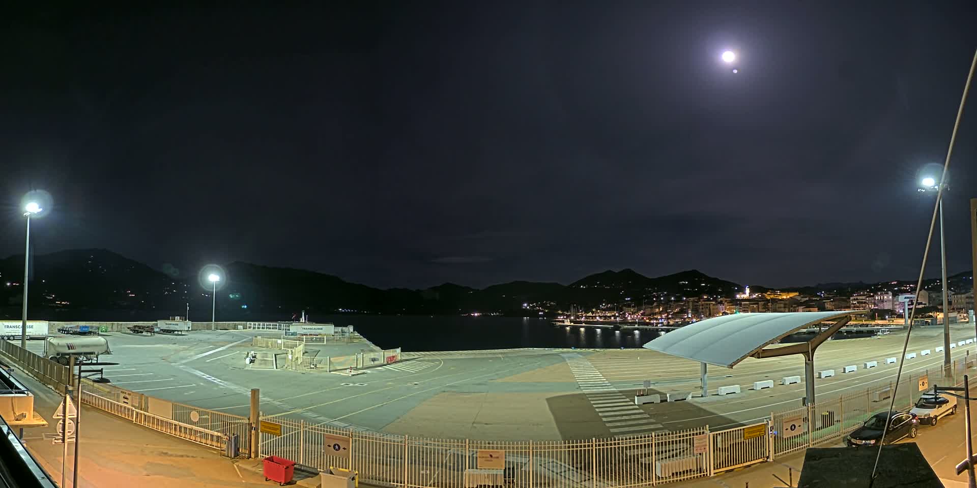 An empty, brightly lit port area at night, under a clear sky with a bright moon, overlooks a dark body of water and distant hills with city lights.