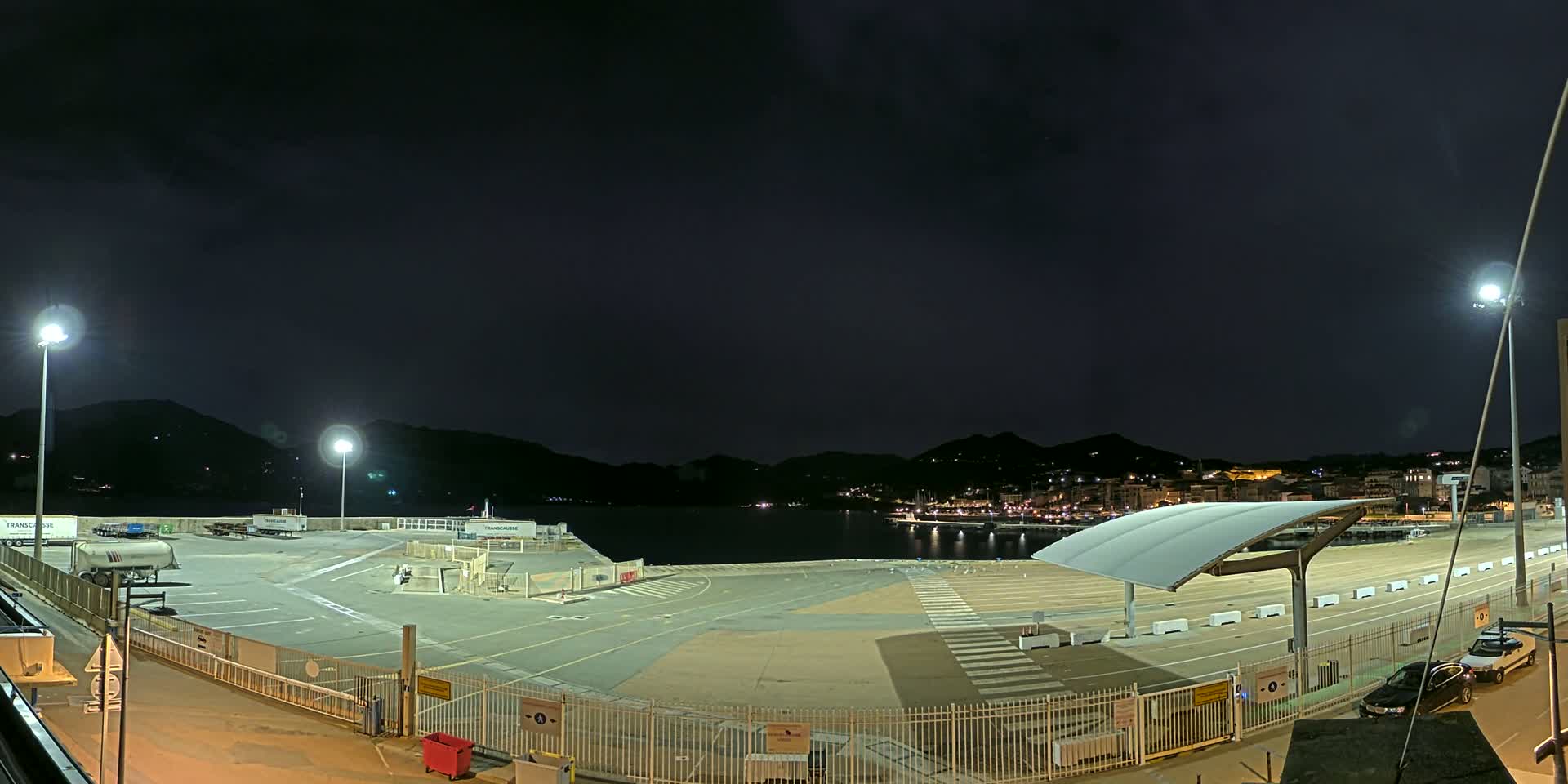 A nighttime view of an empty port area with a few parked vehicles, illuminated by artificial lights, and a dark, calm body of water behind it, with hills and a town visible in the distance under a clear, dark sky.