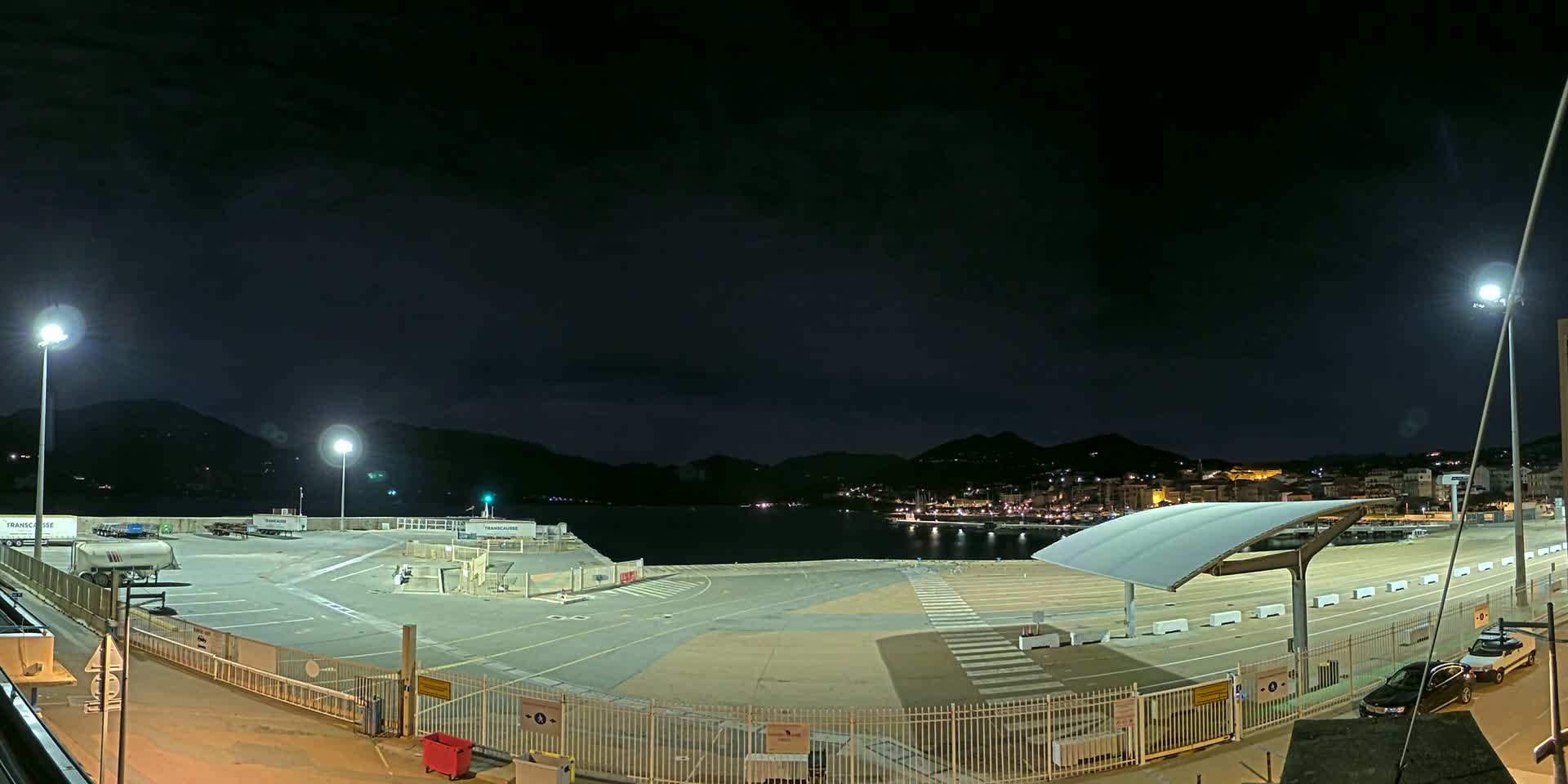 An empty, brightly lit ferry terminal at night overlooks a dark body of water with hills and a town visible in the distance under a cloudy sky.