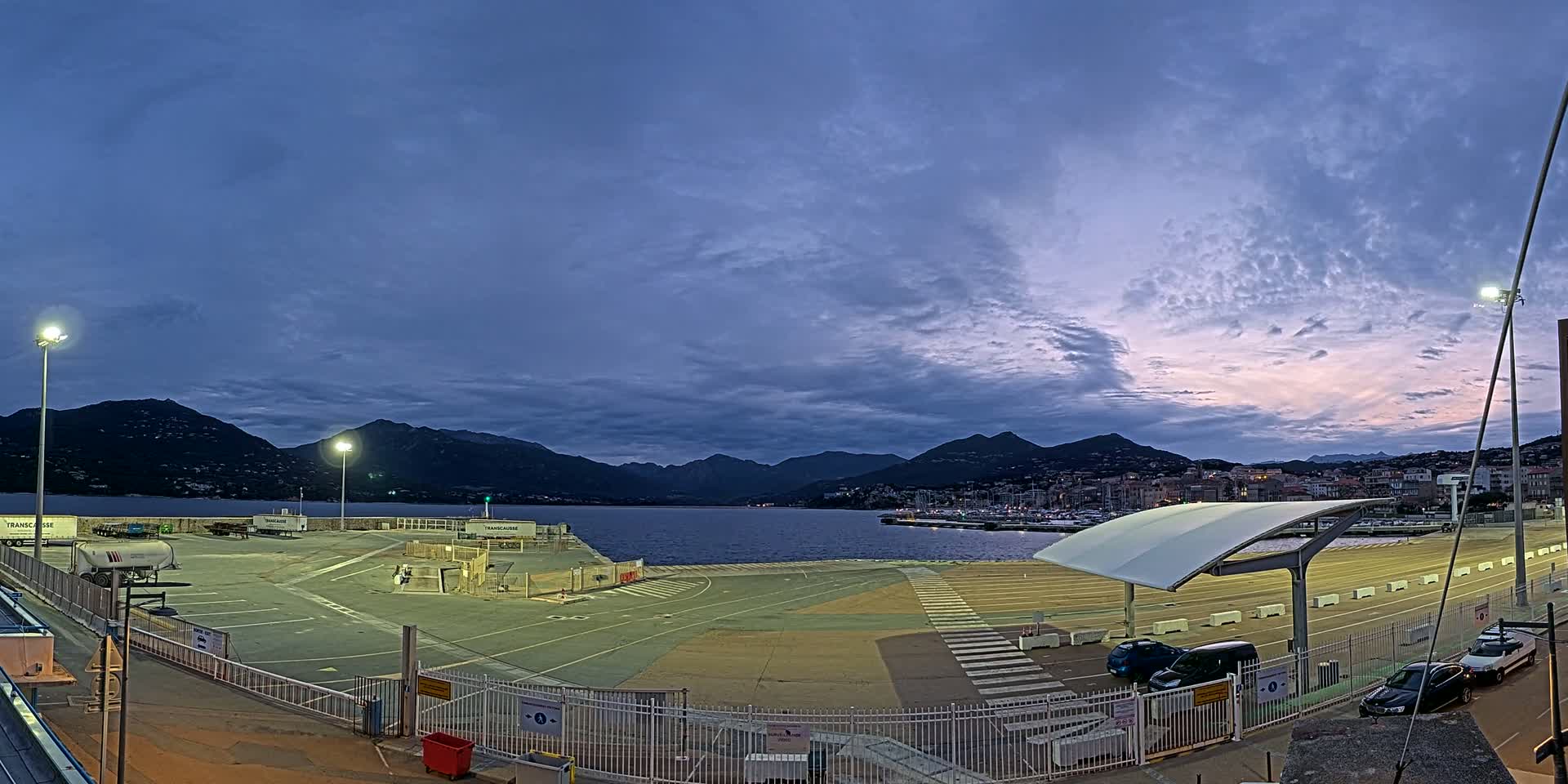 An empty ferry terminal at twilight, under a partly cloudy sky, overlooks a calm body of water and mountains in the distance.